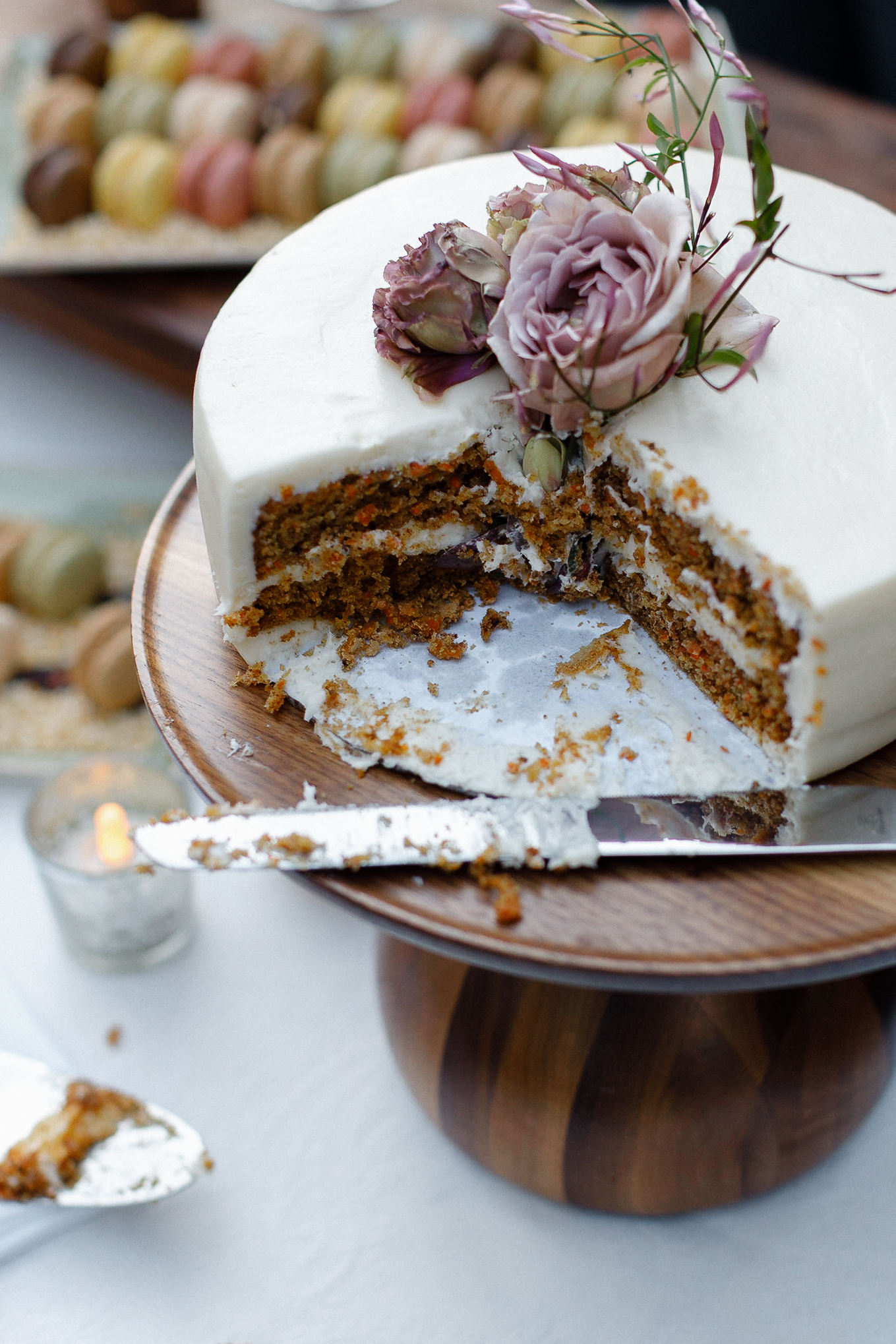 Partially sliced carrot cake with white icing and flowers on a wood stand; knife, plated slice, macarons, and lit candle nearby.