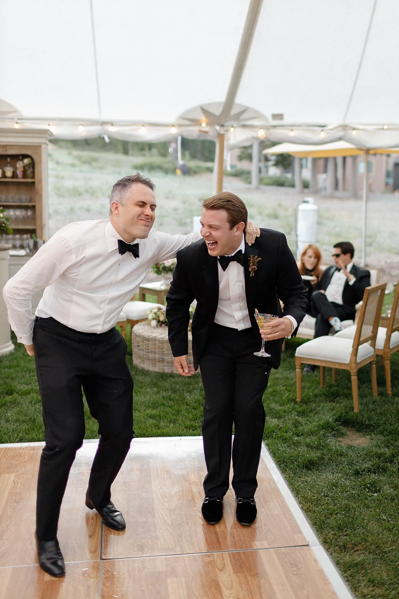 Two men in tuxedos laugh and dance on a wooden floor at a Lake Tahoe event. One holds a drink; guests sit under a lit tent.