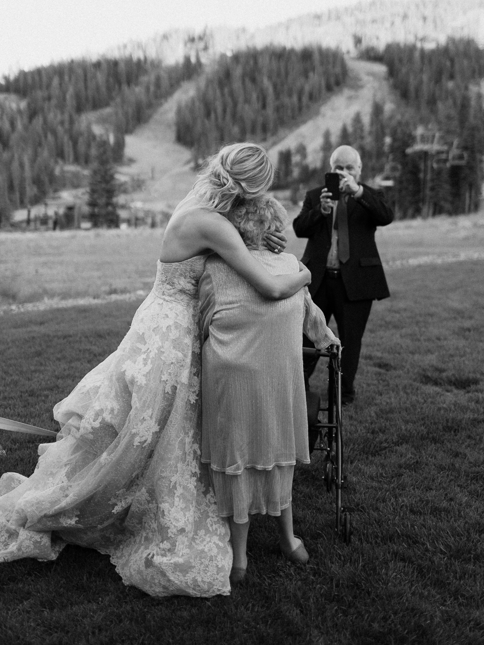 Bride in lace gown hugs elderly woman with walker at outdoor Lake Tahoe wedding; mountains behind, man in suit takes photo.