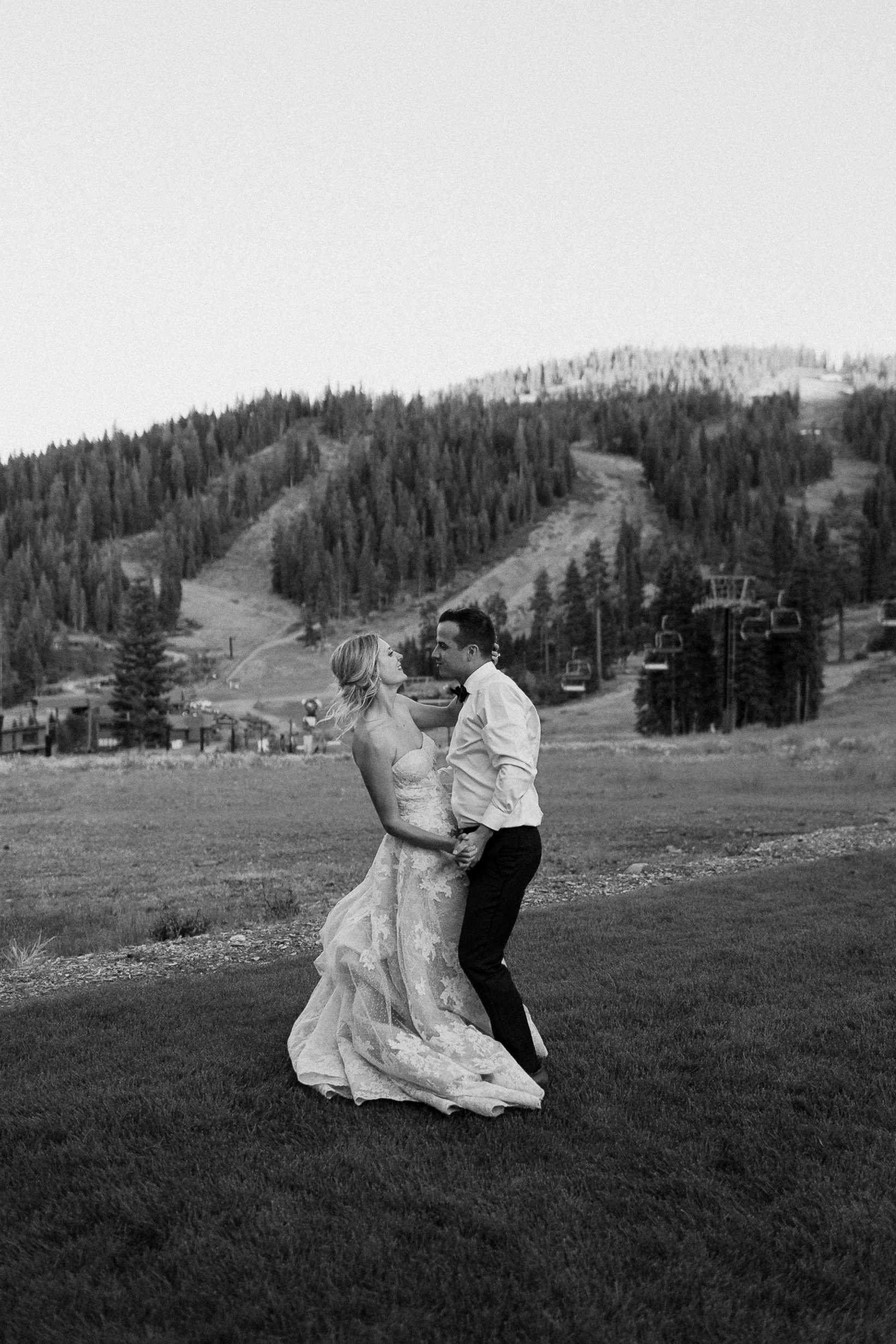 Bride and groom dance on a grassy field with mountains and trees in the black-and-white background, conveying a carefree mood.