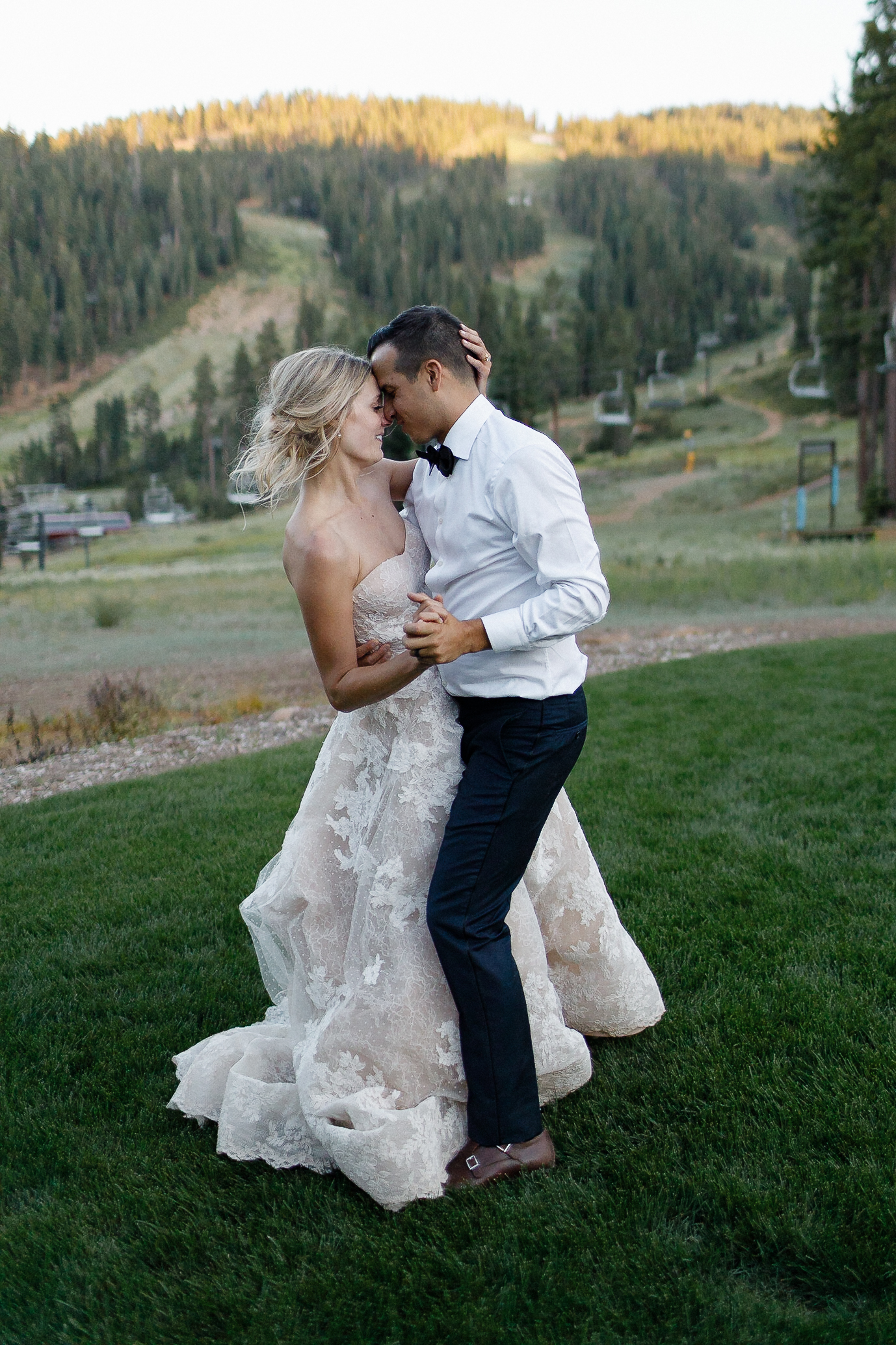Bride in lace gown and groom in white shirt with black bow tie dance on grass, mountains and trees at sunset in the background.