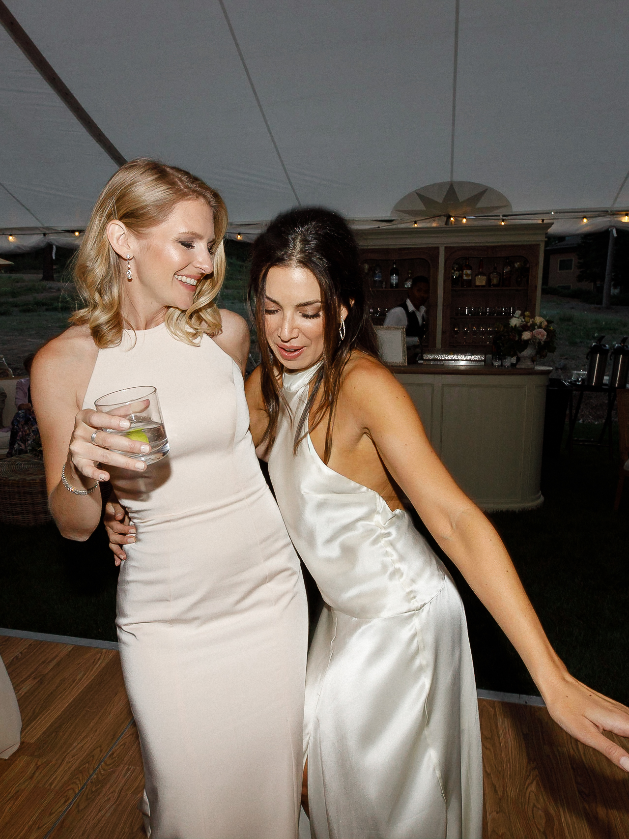 Two women in light dresses smile and dance on a tented wedding floor. One holds a drink, with a floral bar behind at Lake Tahoe.