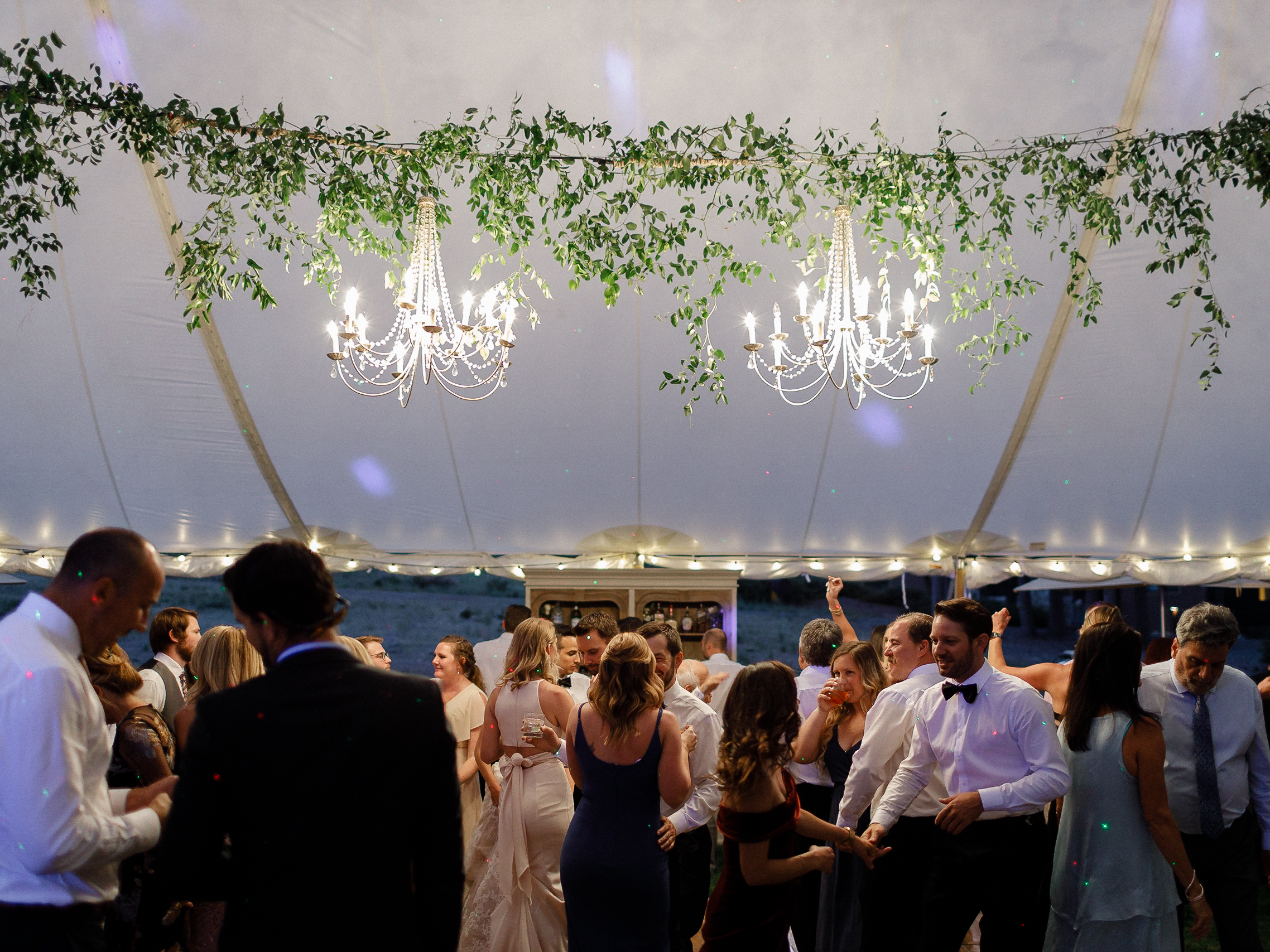 People in formal wear dance and socialize under a white tent with chandeliers and greenery at a festive Lake Tahoe wedding.