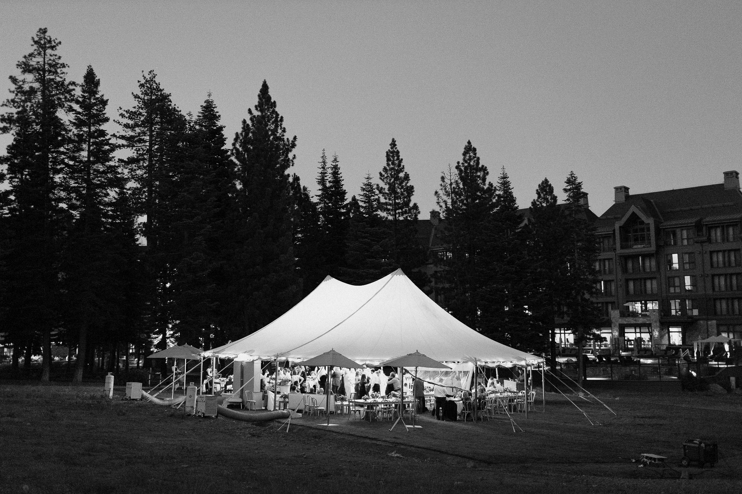 White event tent lit from inside on grass, surrounded by pine trees near Lake Tahoe; people at tables, Ritz-Carlton in background.