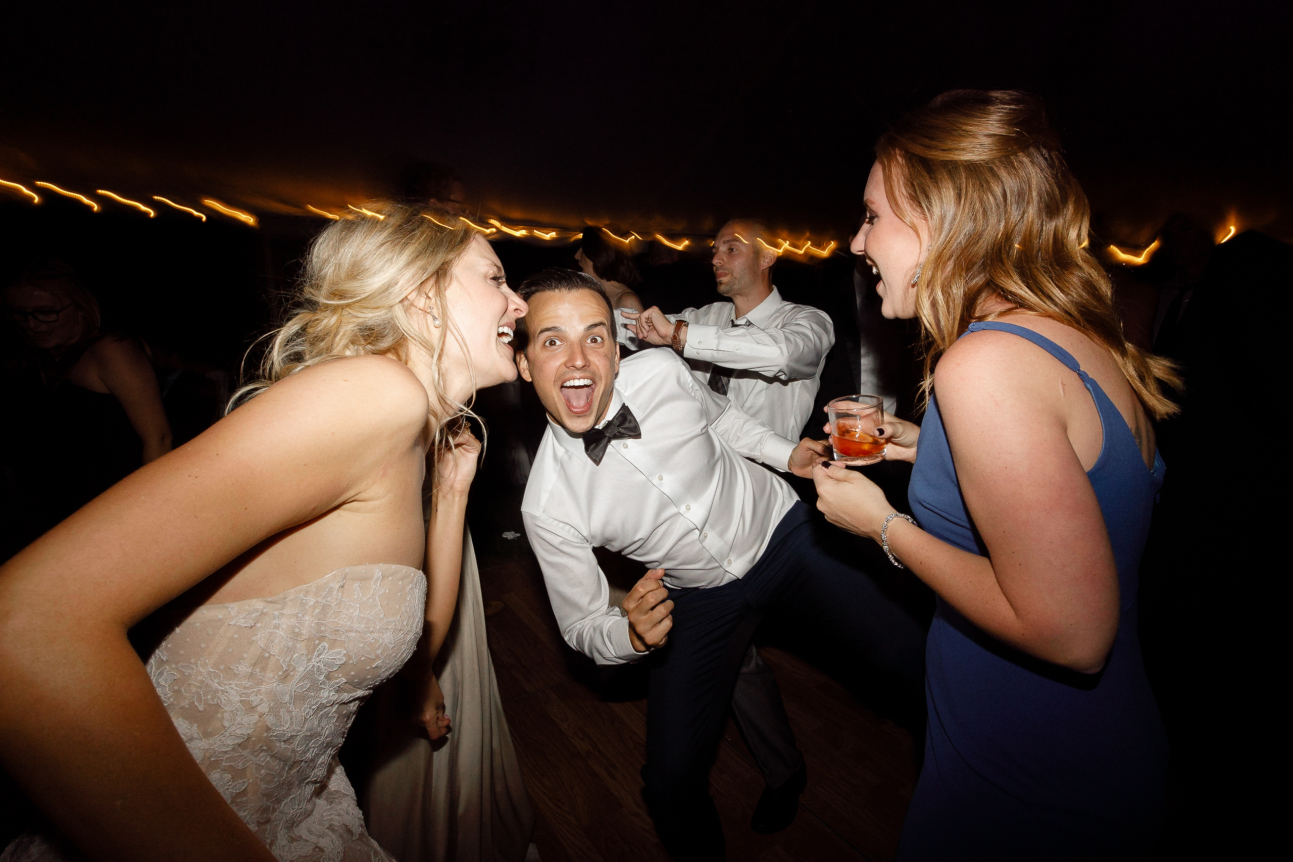 Three formally dressed people laugh and dance at a Lake Tahoe wedding, holding drinks, with string lights in the background.