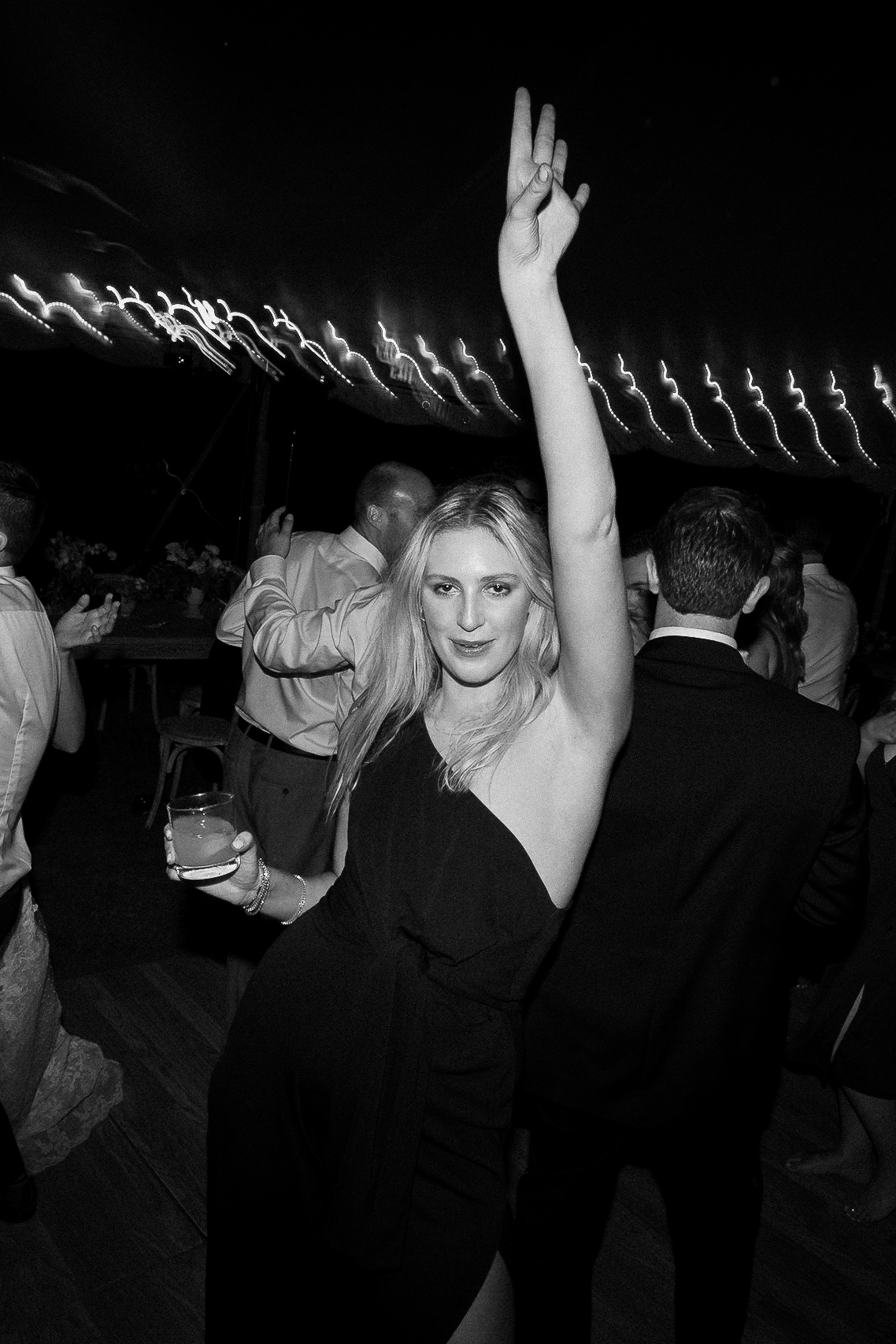 Woman in black dress poses, one arm up, holding a drink. Background: guests in formal wear dance under string lights at wedding.