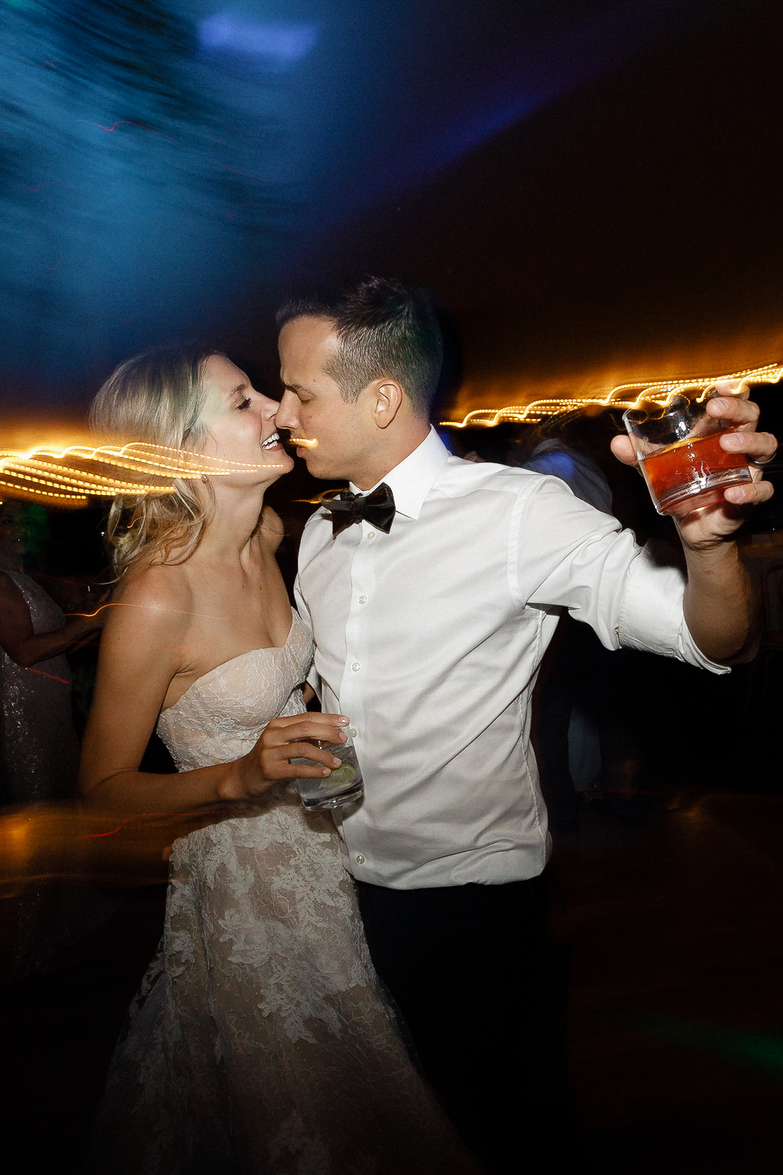 Bride and groom smile and embrace on the Ritz-Carlton dance floor at night, holding drinks under string lights at their wedding.