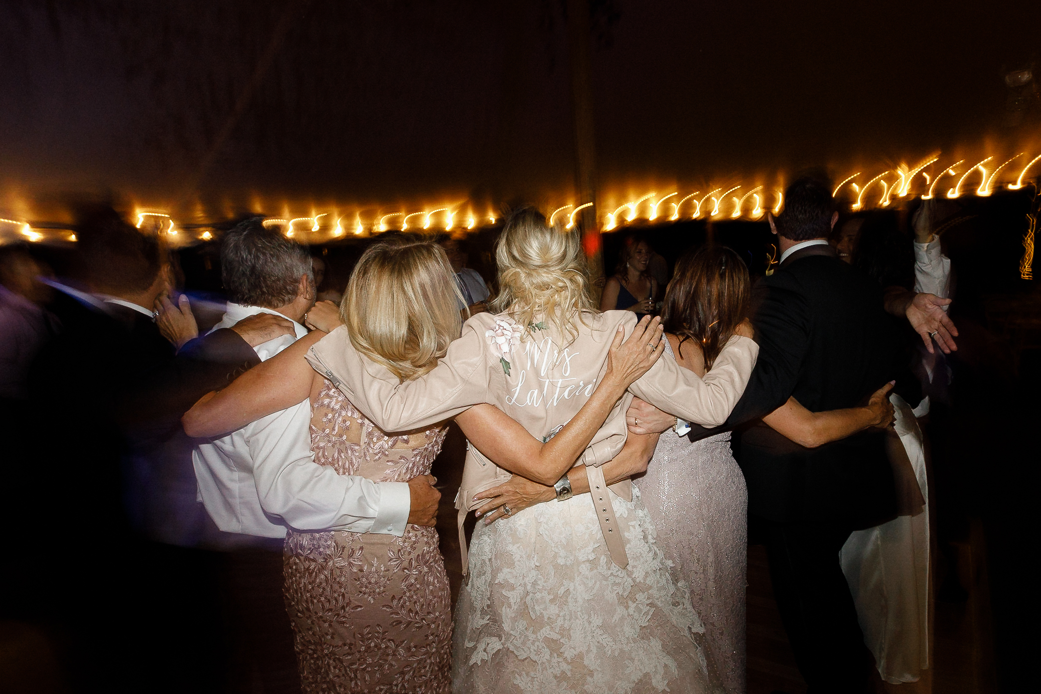 Wedding guests in formal attire, including a bride in a "Mrs" jacket, stand arm-in-arm under string lights at Ritz-Carlton Lake Tahoe.