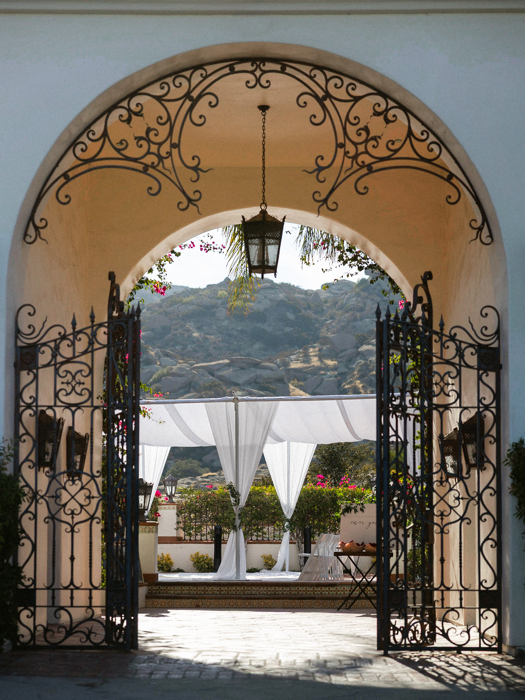 Wrought iron gates open to an arched entry with lantern, leading to a white canopy with drapes and mountain views at Hummingbird Nest Events.