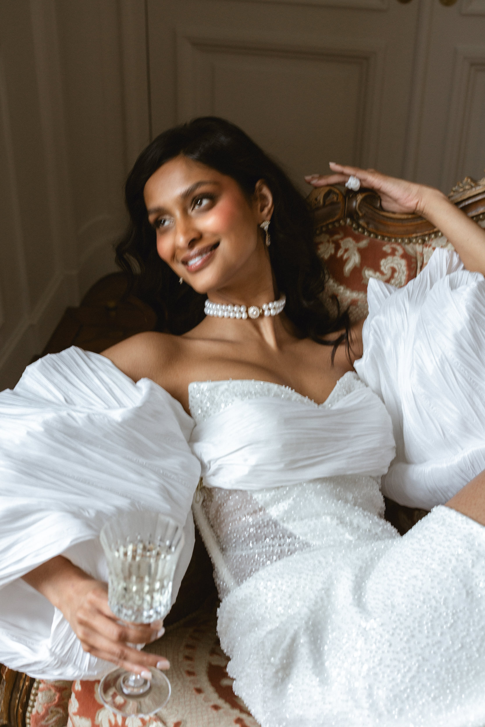 Woman in white wedding gown with pleated sleeves reclines on ornate chair, smiling and holding a crystal glass; wears pearl choker.