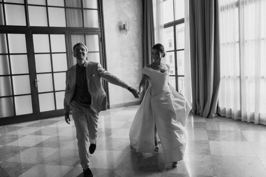 A smiling wedding couple in formal attire walk hand in hand on a checkered marble floor in Sibenik, natural light filling the room.