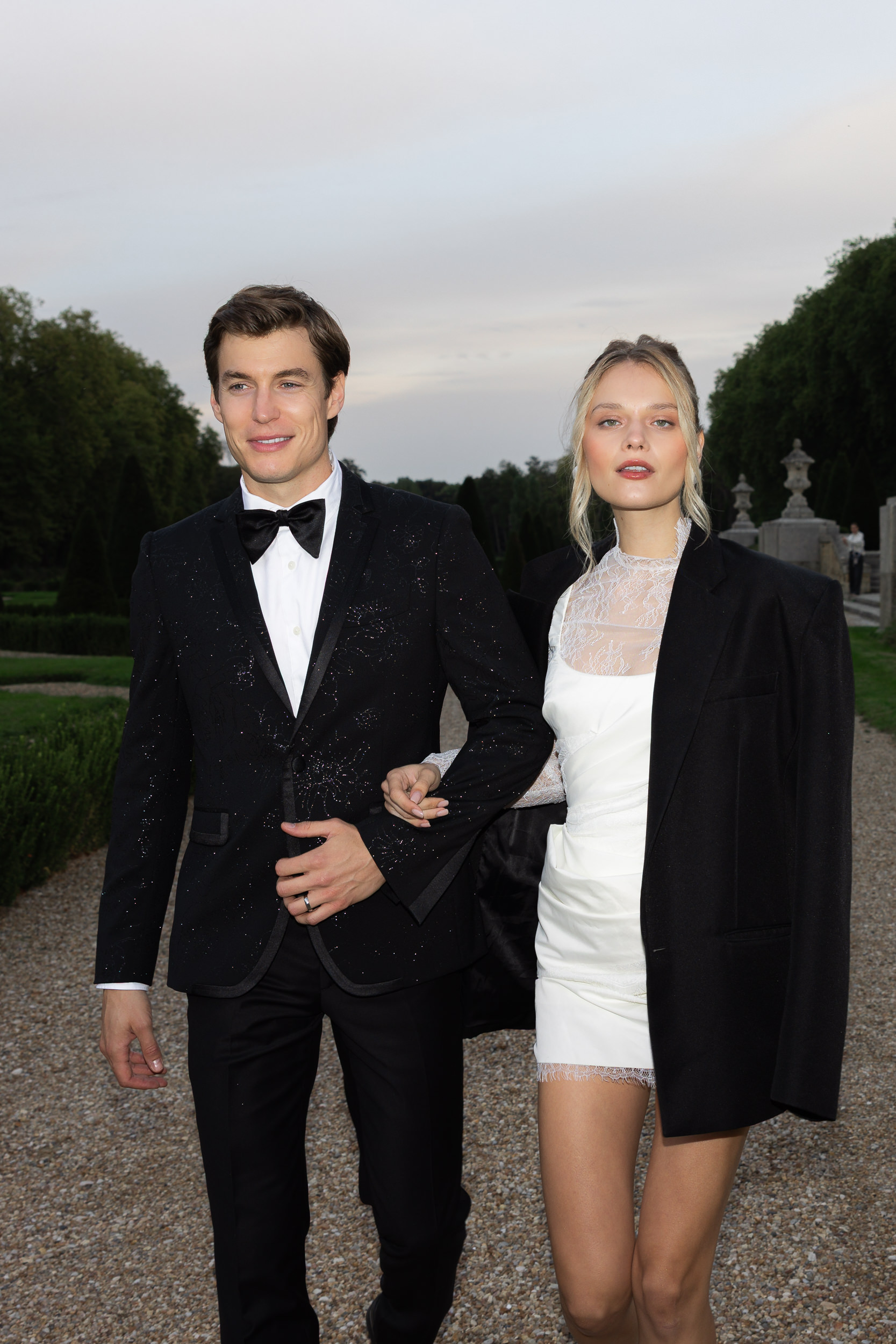 A man in a black tuxedo and a woman in a white dress with a black jacket walk arm in arm on a gravel path at Chateau de Villette.