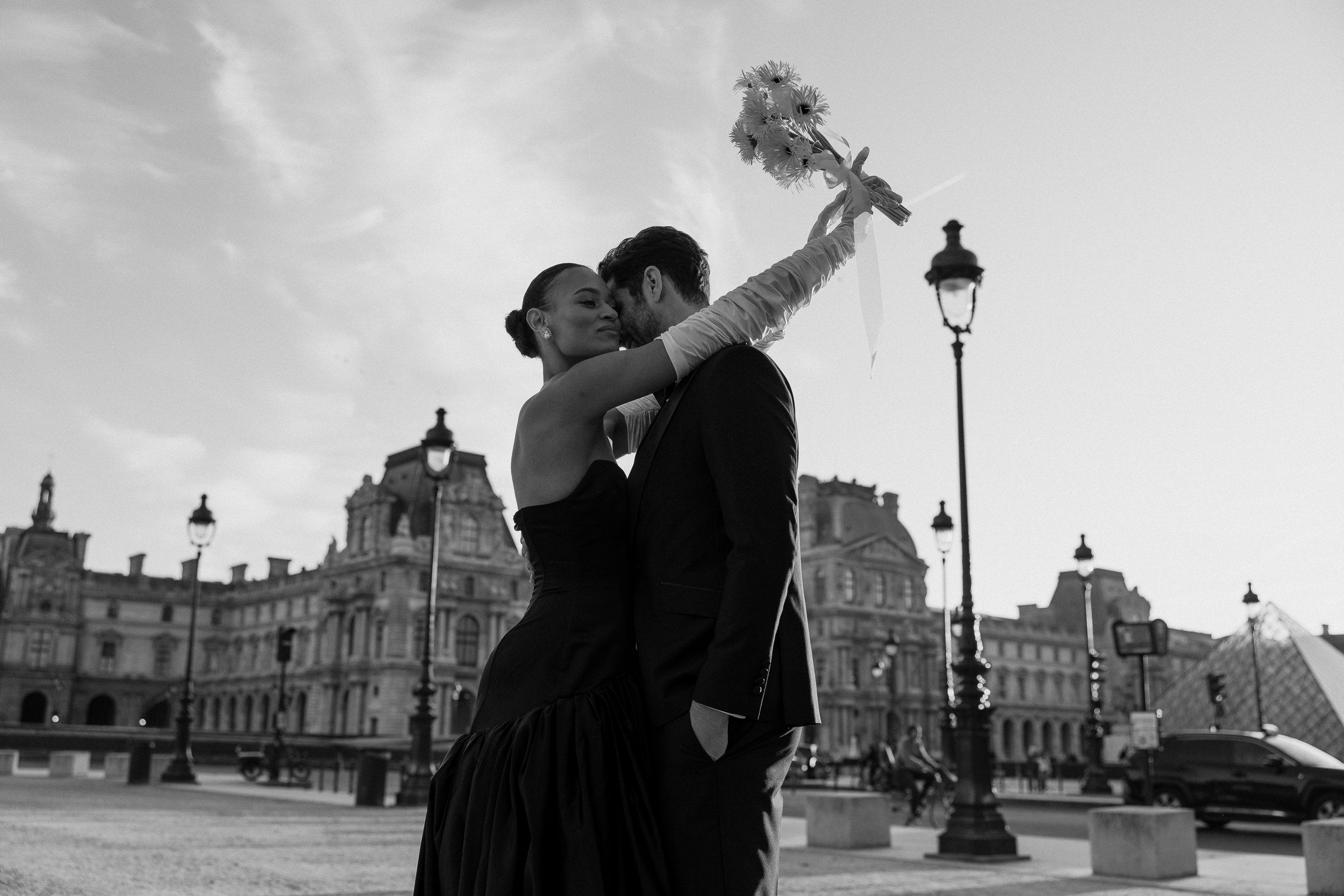 A woman in a dark dress hugs a man in a suit, holding up a bouquet near Parisian buildings and street lamps by the Louvre.