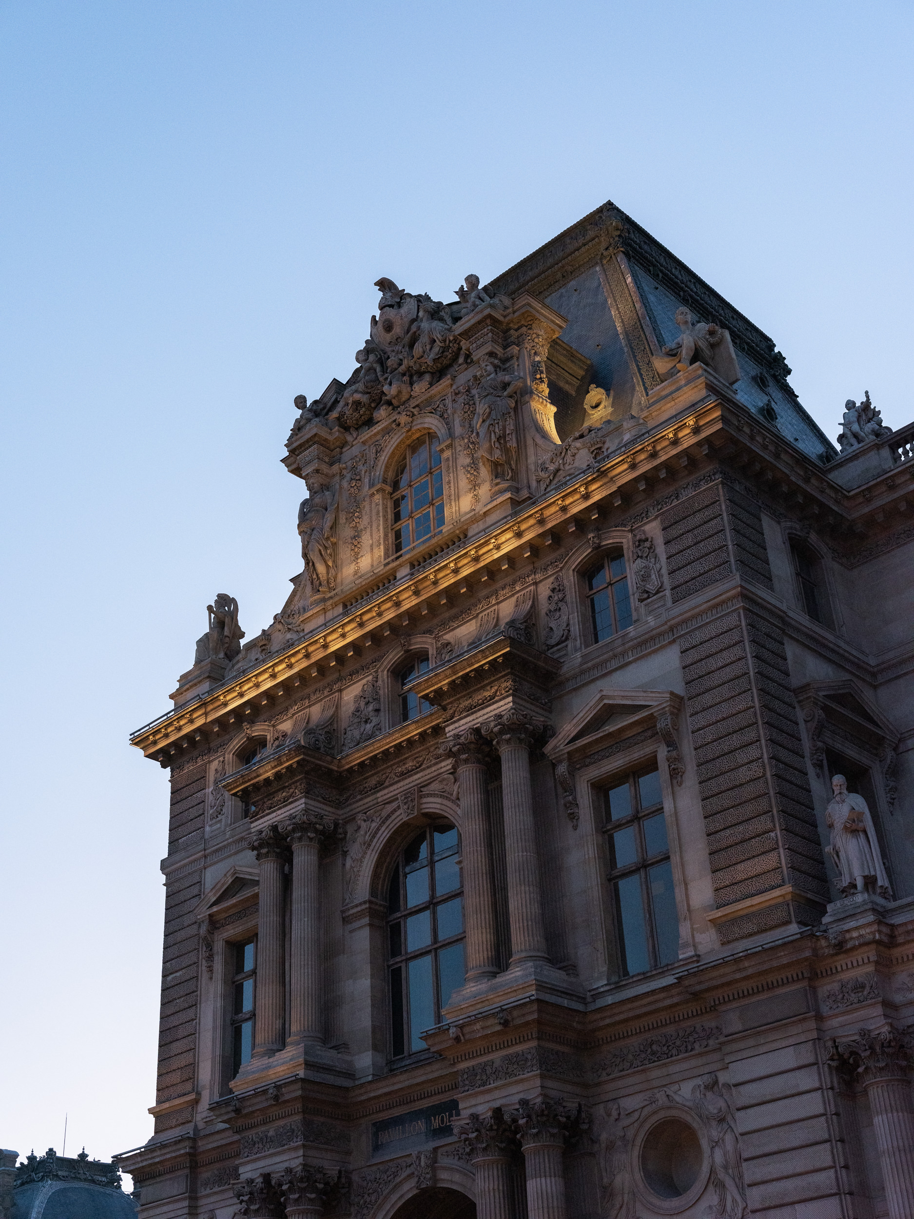 Ornate stone façade of the Louvre in Paris with sculptures, columns, and windows, lit by evening light against a clear blue sky.