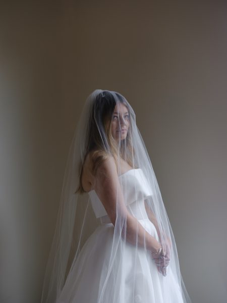 A bride in a white dress with a long sheer veil stands at Cavallo Point, hands clasped, facing forward against a neutral backdrop.