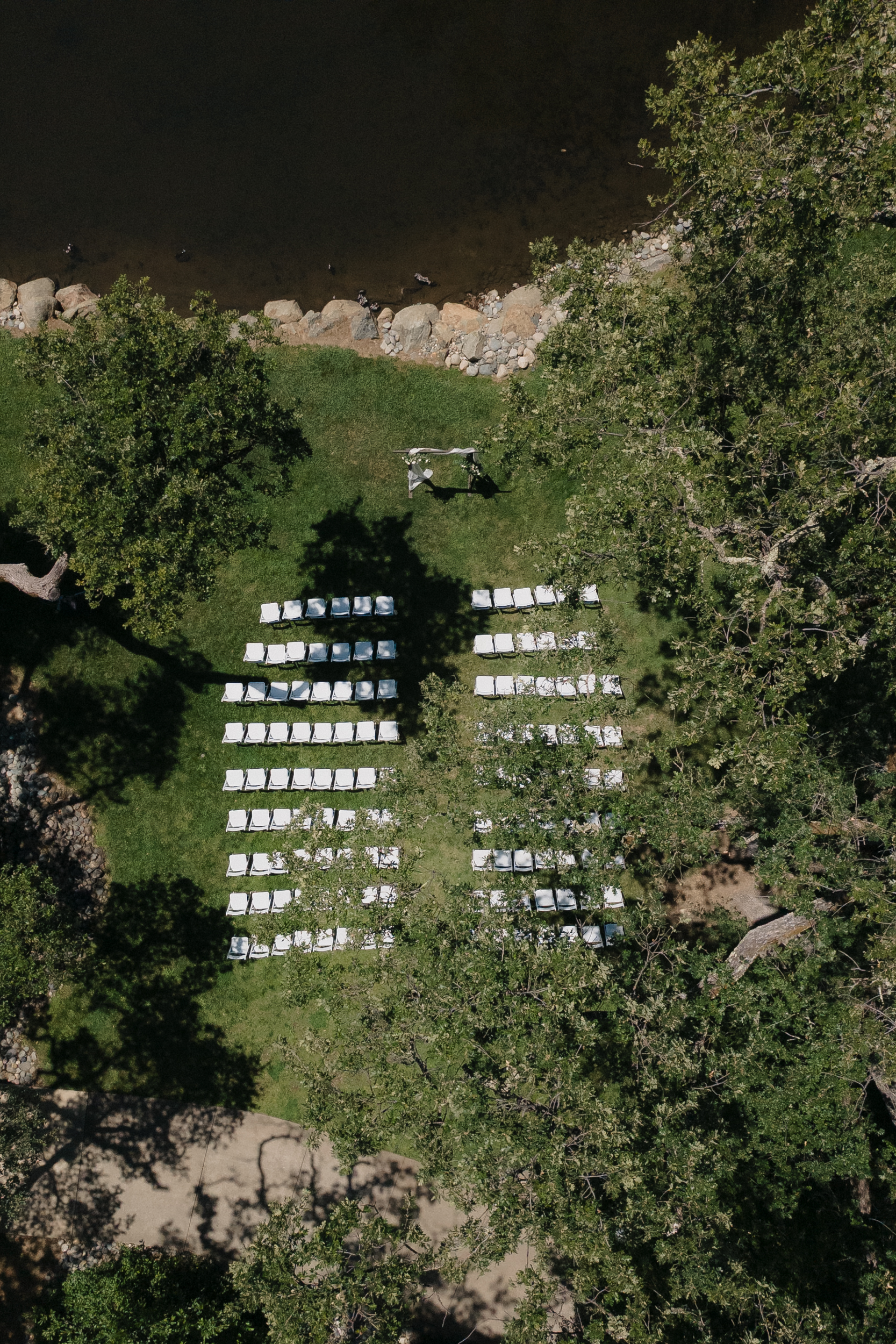 Aerial view of a California outdoor wedding setup: white chairs on grass facing an arch, trees, and a rocky shoreline nearby.