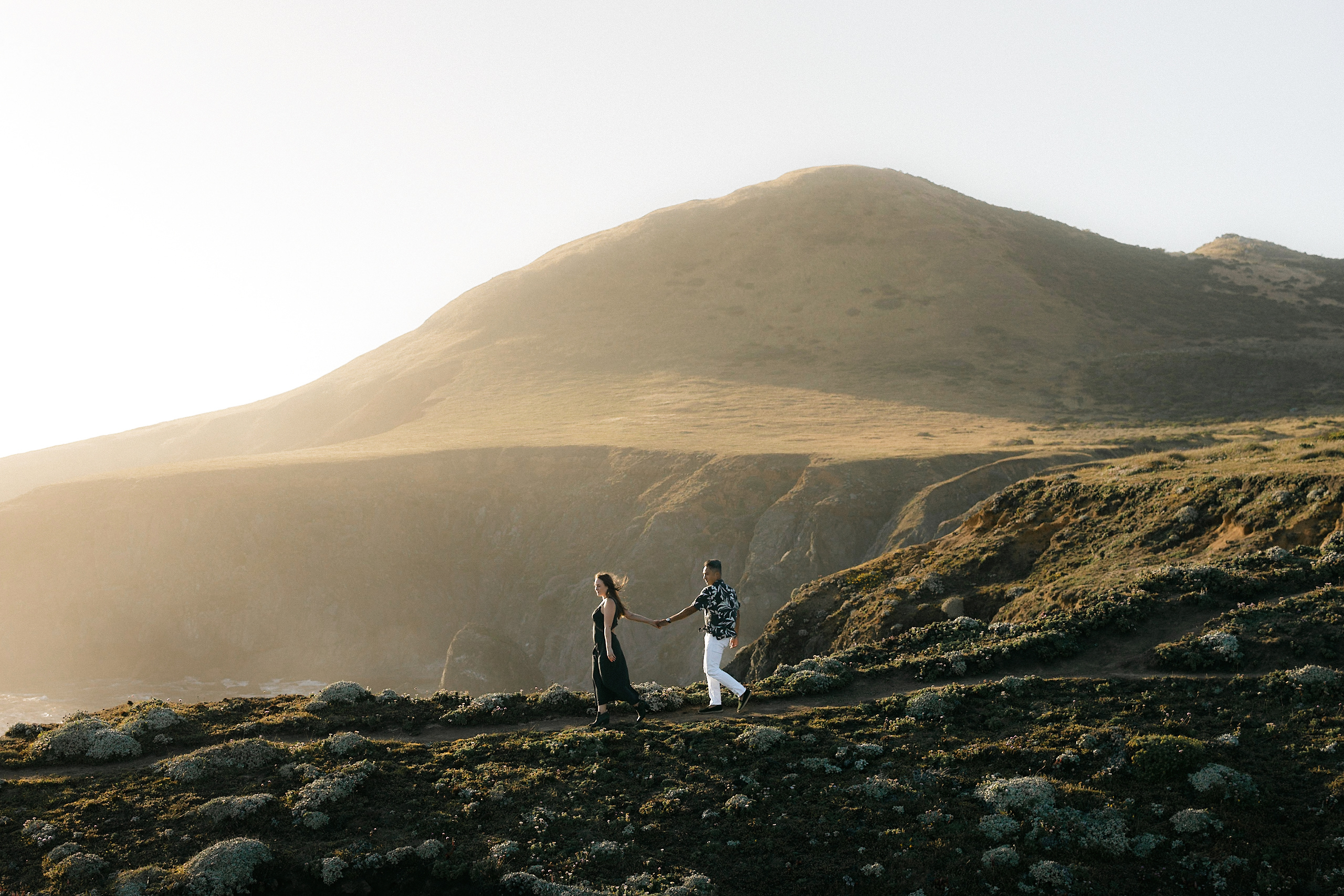 Elegant couple holds hands on a cliff, golden light, serene San Francisco engagement photo.