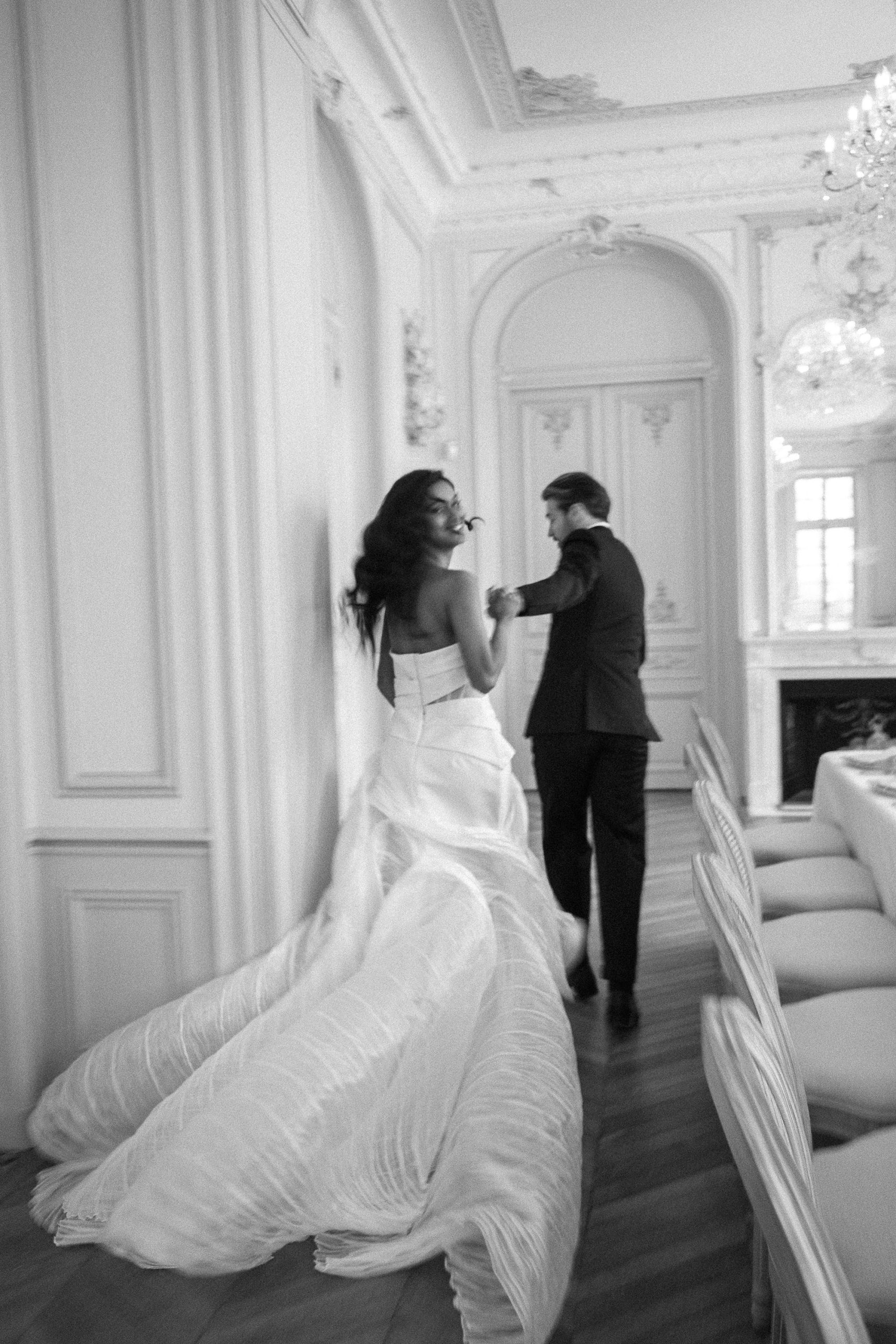 Bride and groom hold hands, walking through an ornate Chateau Nainville les Roches room with chandeliers. Black and white photo.