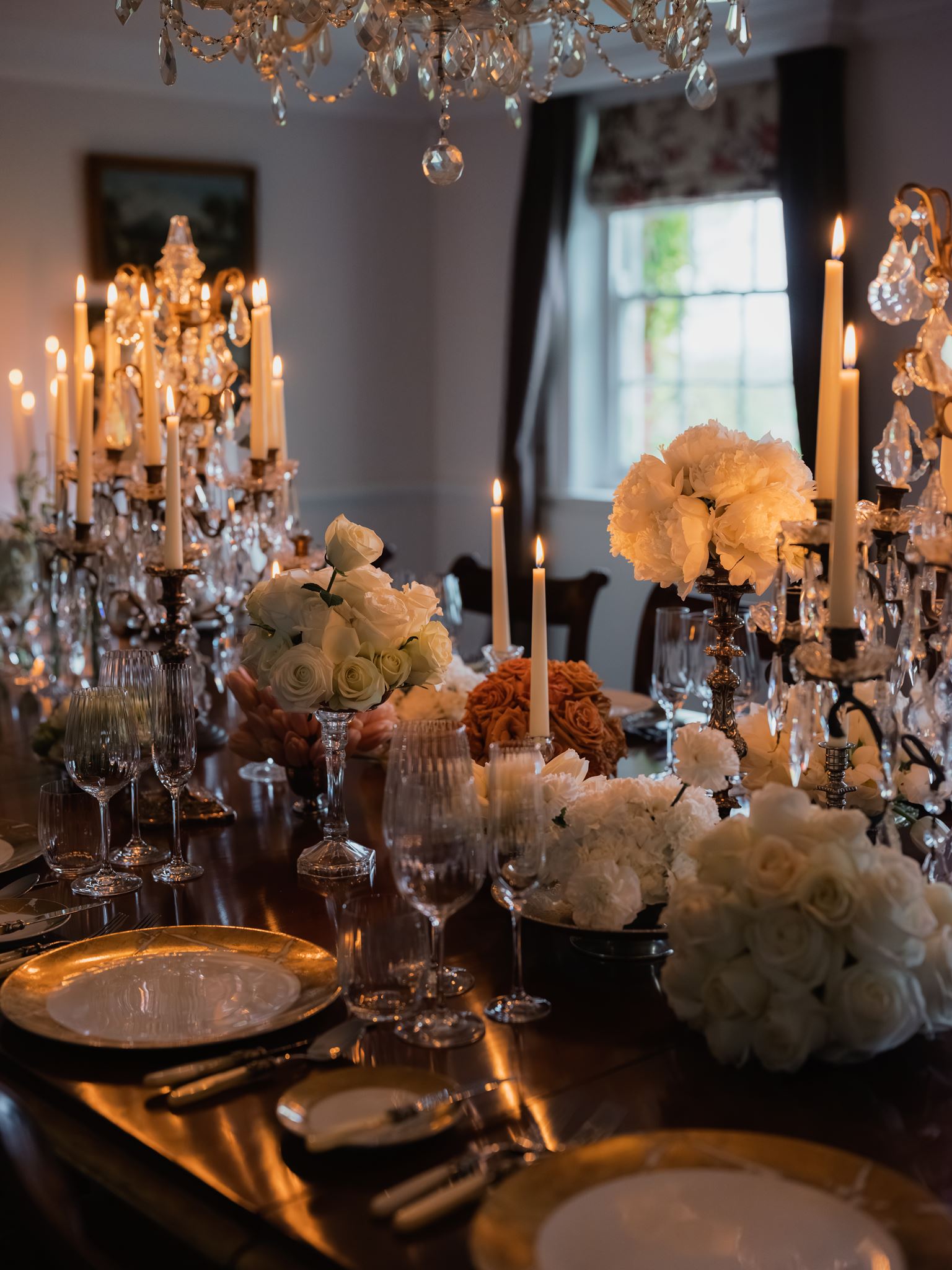 A dining table with gold-rimmed plates, crystal glasses, and pink flowers in a softly lit room; the couple's car outside the window.