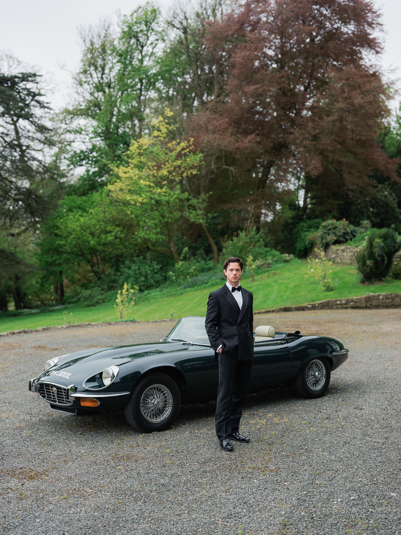 A man in a black tuxedo stands by a classic dark green convertible on a gravel drive, with green trees and grass in the background.