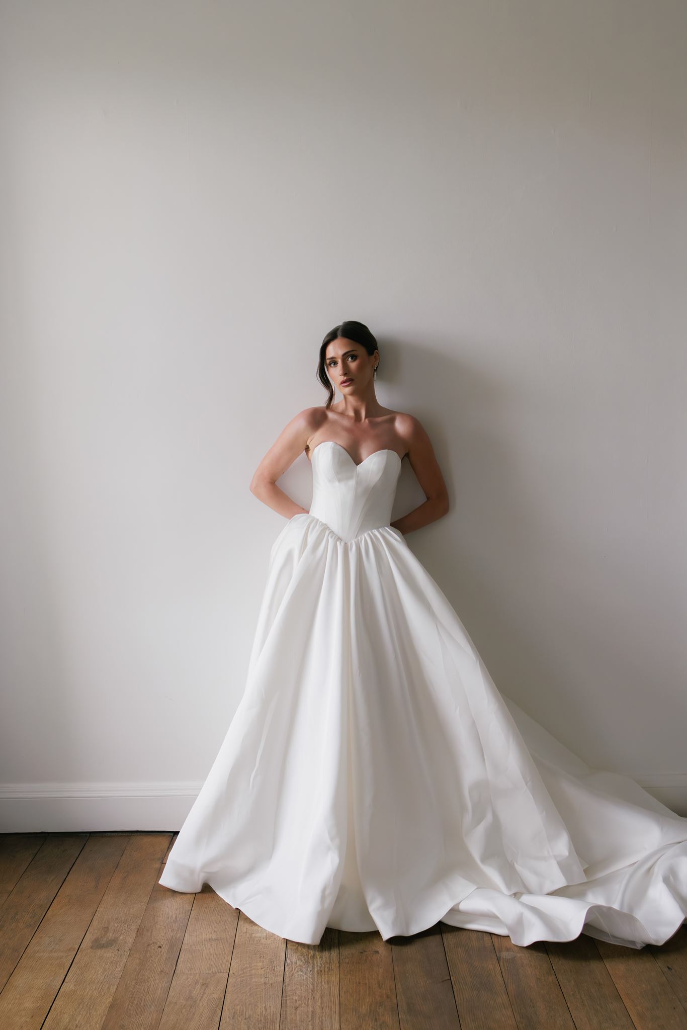 Woman in strapless white wedding gown with full skirt stands against plain wall on wood floor, hands in pockets, looking aside.