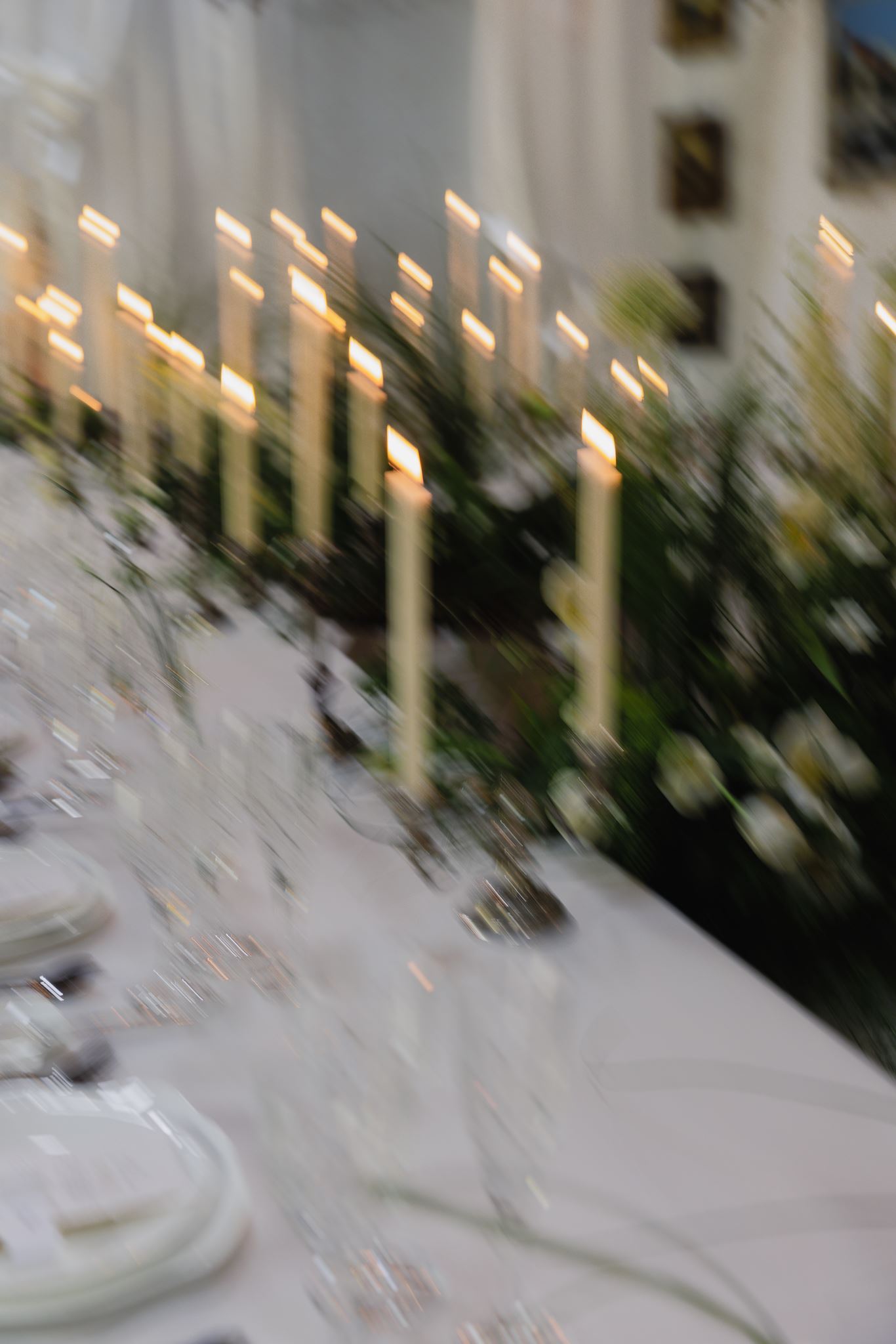 A long table set with white plates, glassware, lit candles, greenery, and white flowers; image has a soft, romantic blur.
