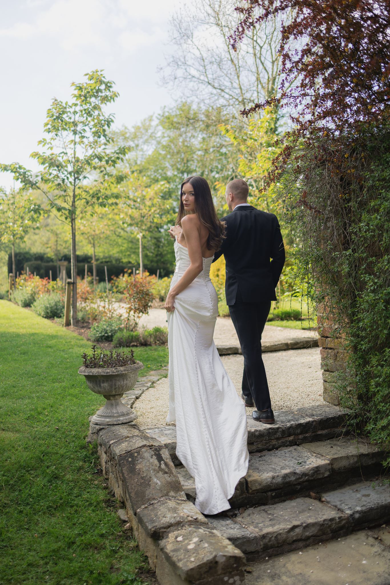 Bride in white dress smiles on stone steps outdoors as groom in black suit walks ahead on a garden path surrounded by greenery.