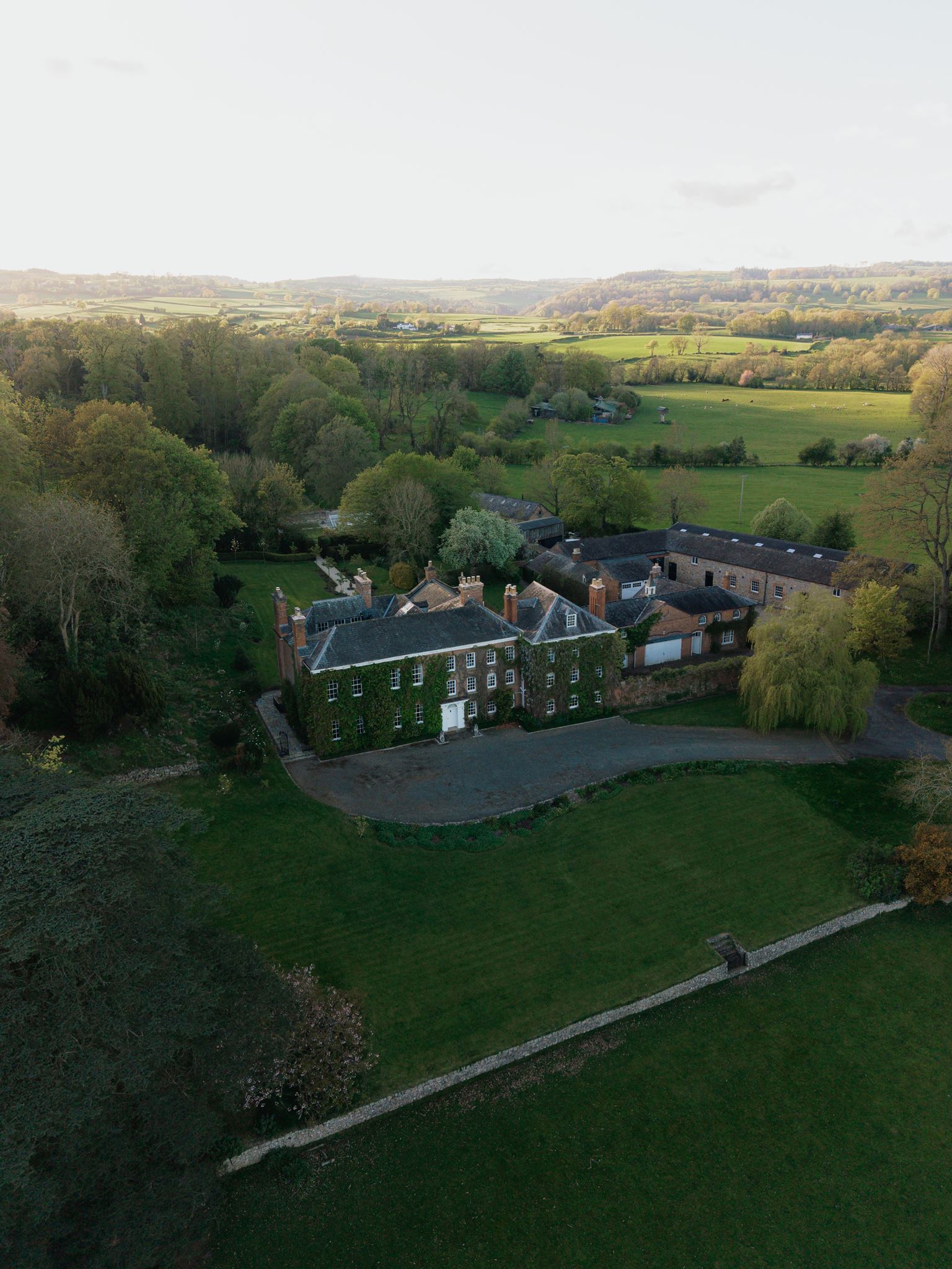 Large ivy-covered manor with several chimneys, green lawns, trees, outbuildings, and fields under a clear sky; fits weddings.