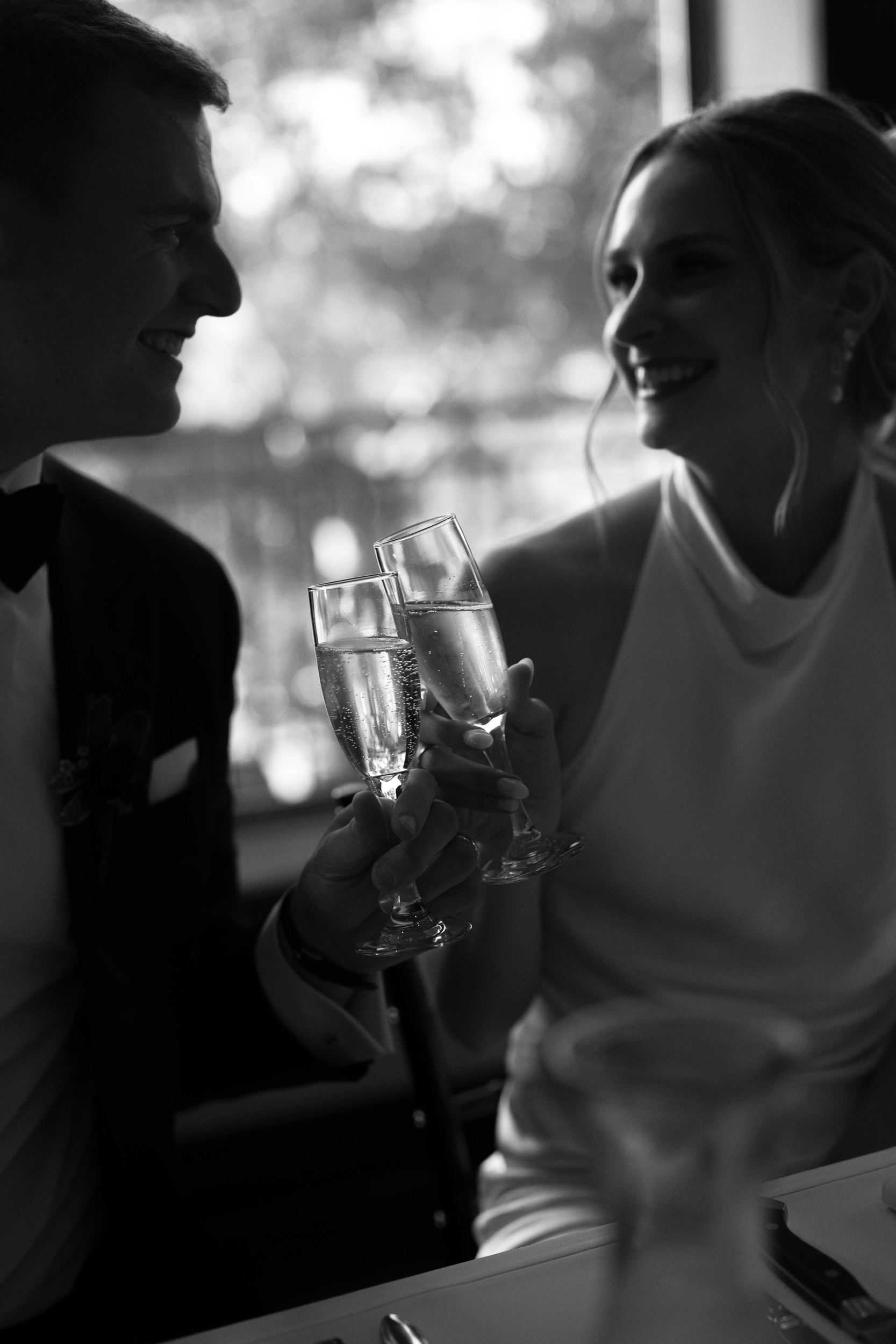 Black and white photo of a bride and groom smiling at each other, clinking champagne glasses at a table by a sunlit window.