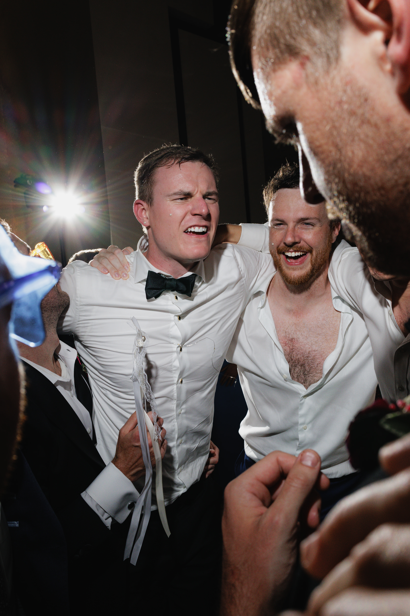 Men in white shirts celebrate at a San Francisco wedding. The joyful groom, in a bow tie, hugs friends as bright light shines behind.