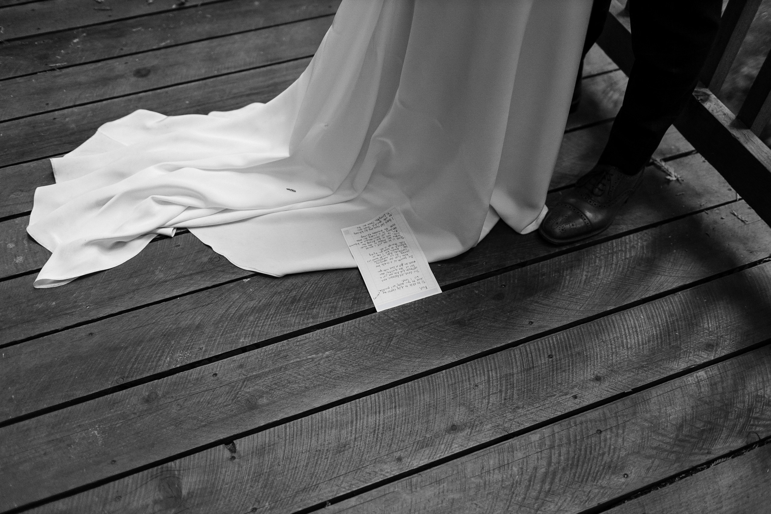 Black & white: Brides flowing train, handwritten notes, grooms polished shoes on wooden floor.