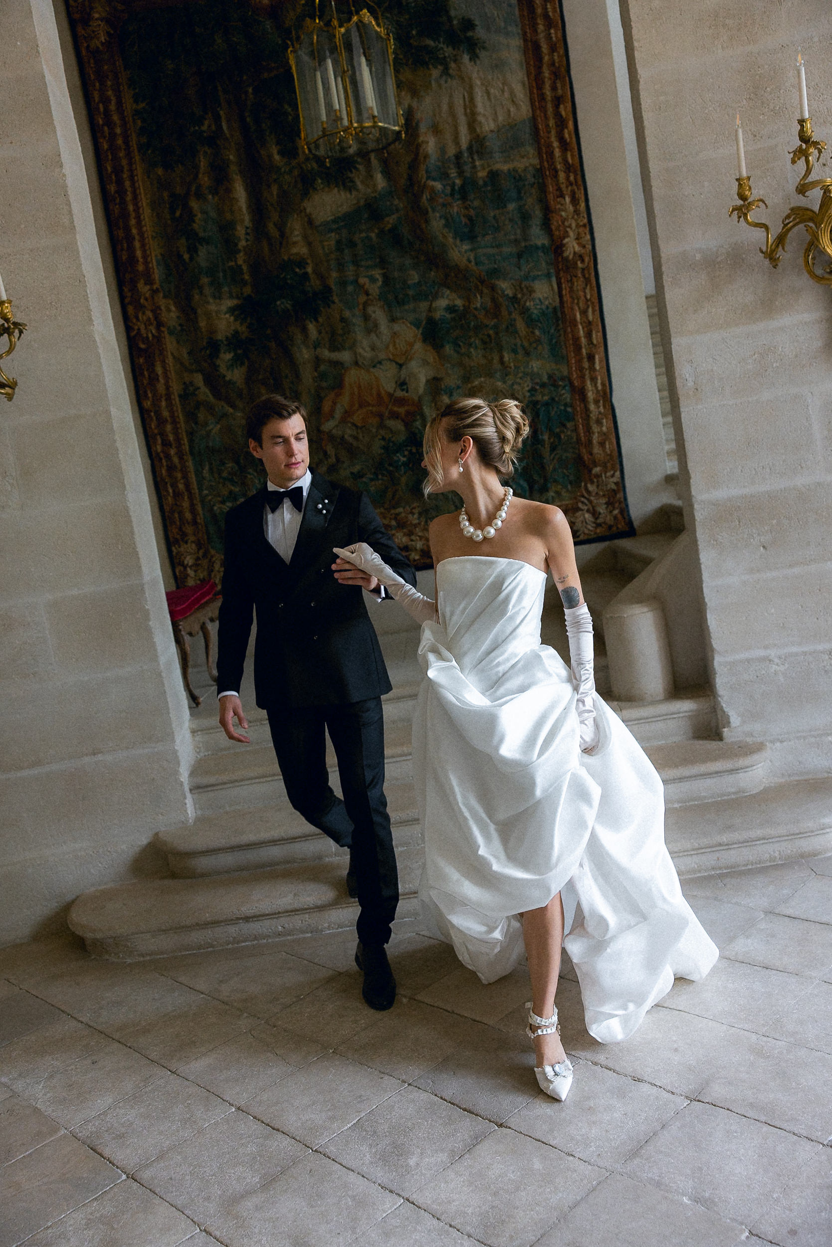 Bride in strapless white gown and partner in black tux walk inside stone-walled chateau de Villette with tapestry and sconces.