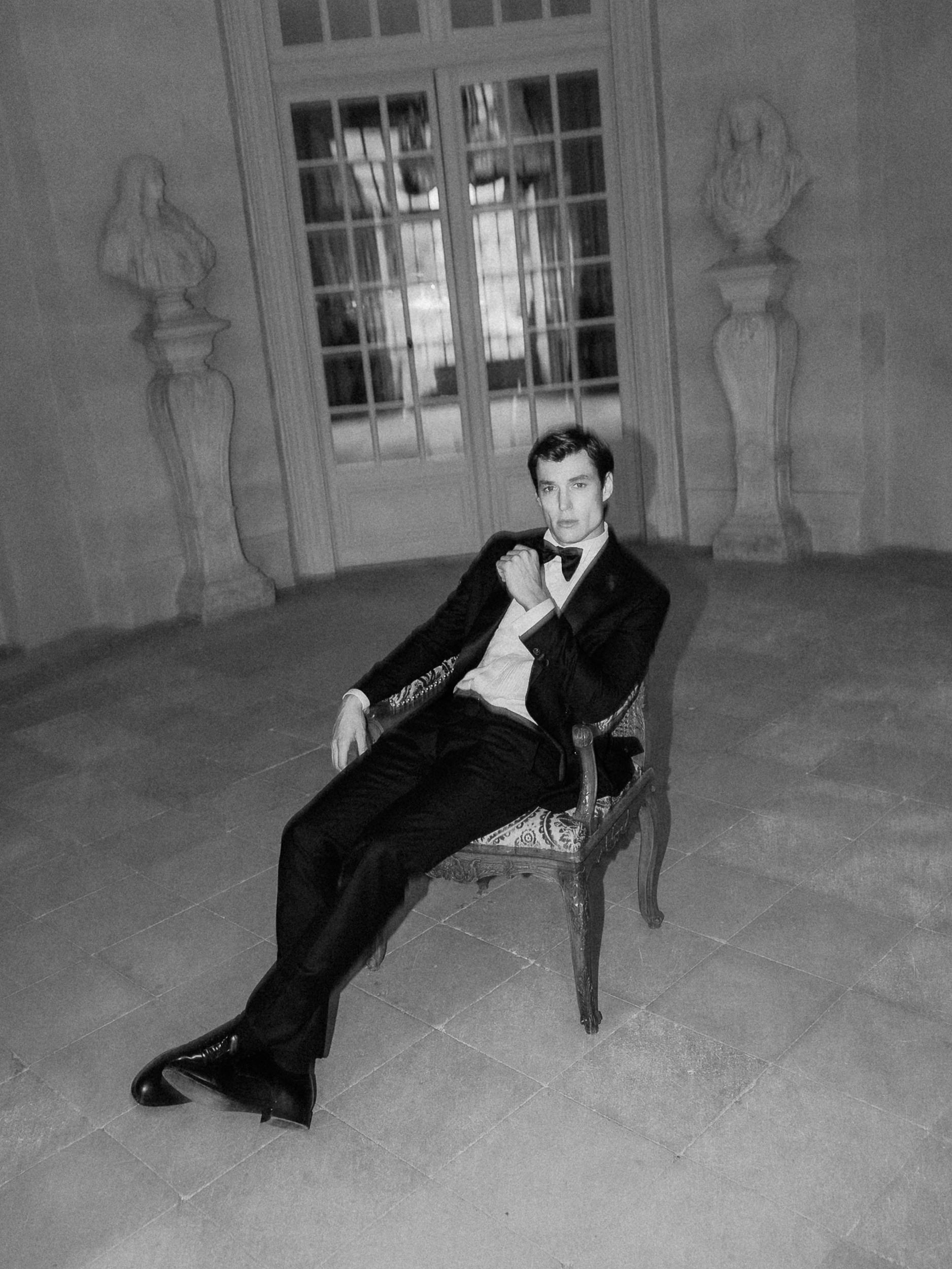 A groom in a tuxedo sits in an ornate chair at Chateau de Villette, surrounded by statues and windows in a classic black and white photo.