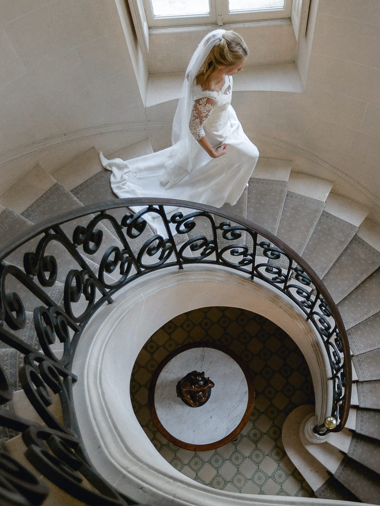 Bride in white dress and veil walks up curved staircase with ornate black railing at Château du Clair de Lune, sunlight above.
