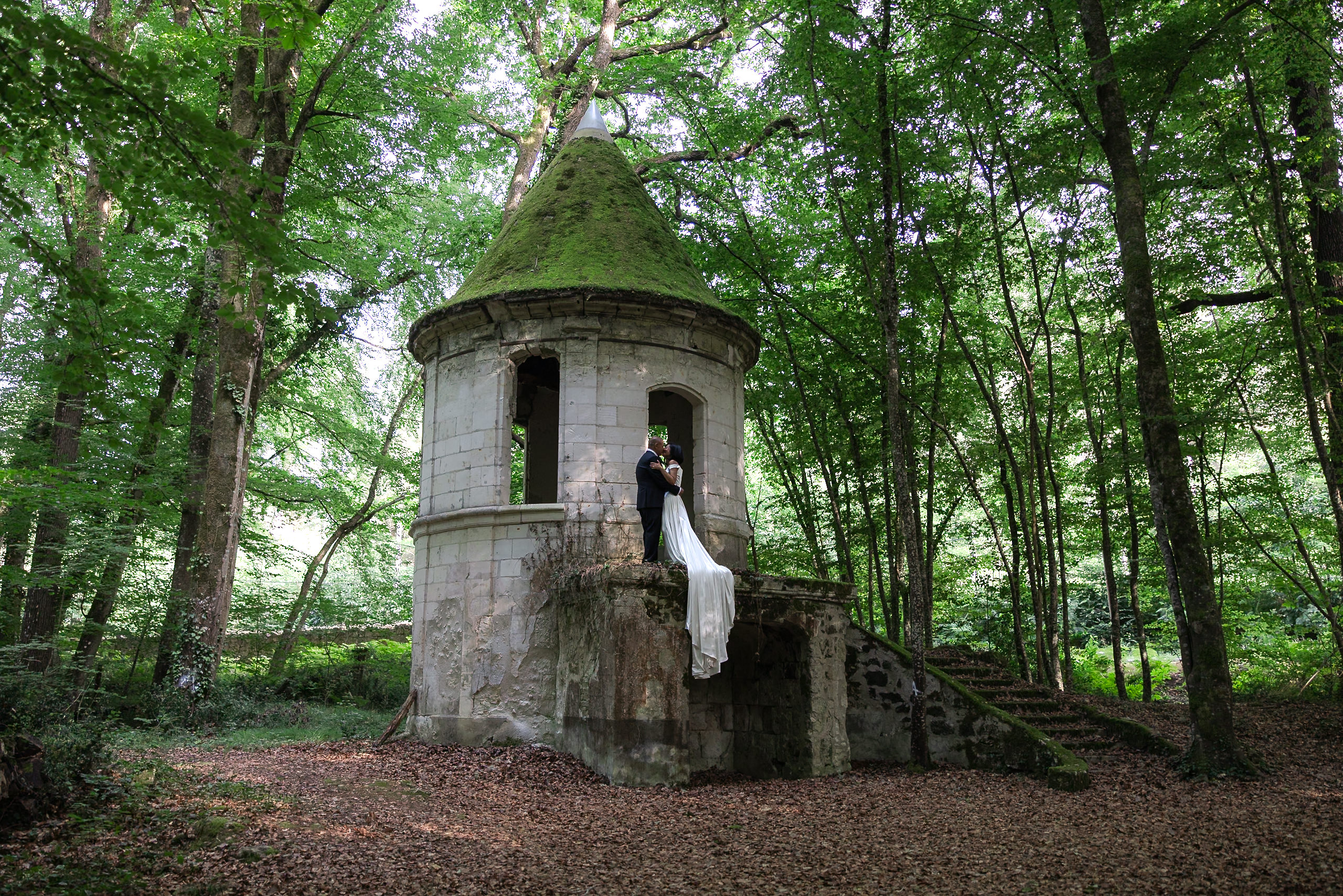 Bride and groom embrace on mossy stone steps at Chateau de Jalesnes; her white dress trails down, leaves scattered around.