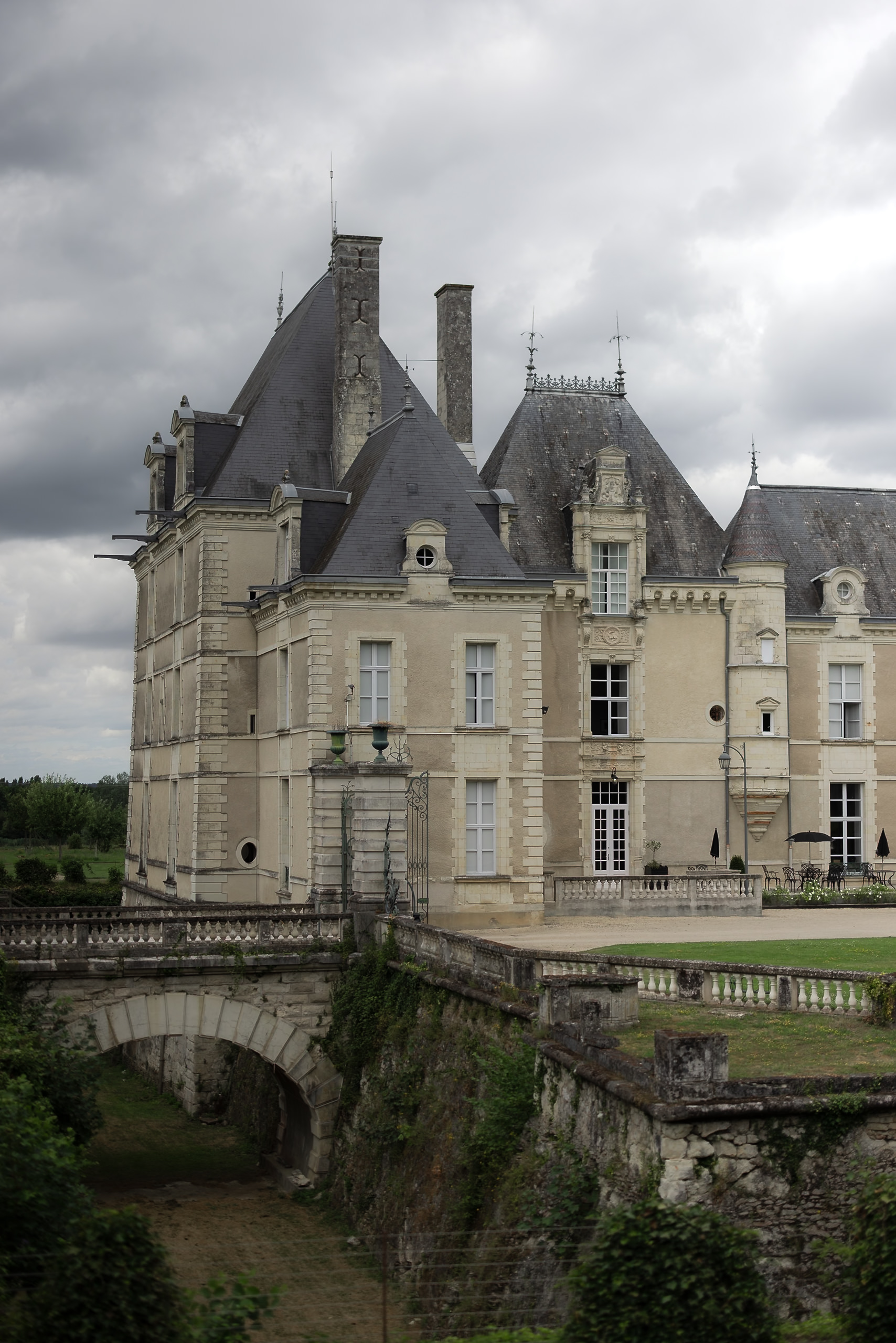 Light stone chateau with tall slate roofs and ornate details beside a stone bridge over a dry moat in Saumur under cloudy skies.