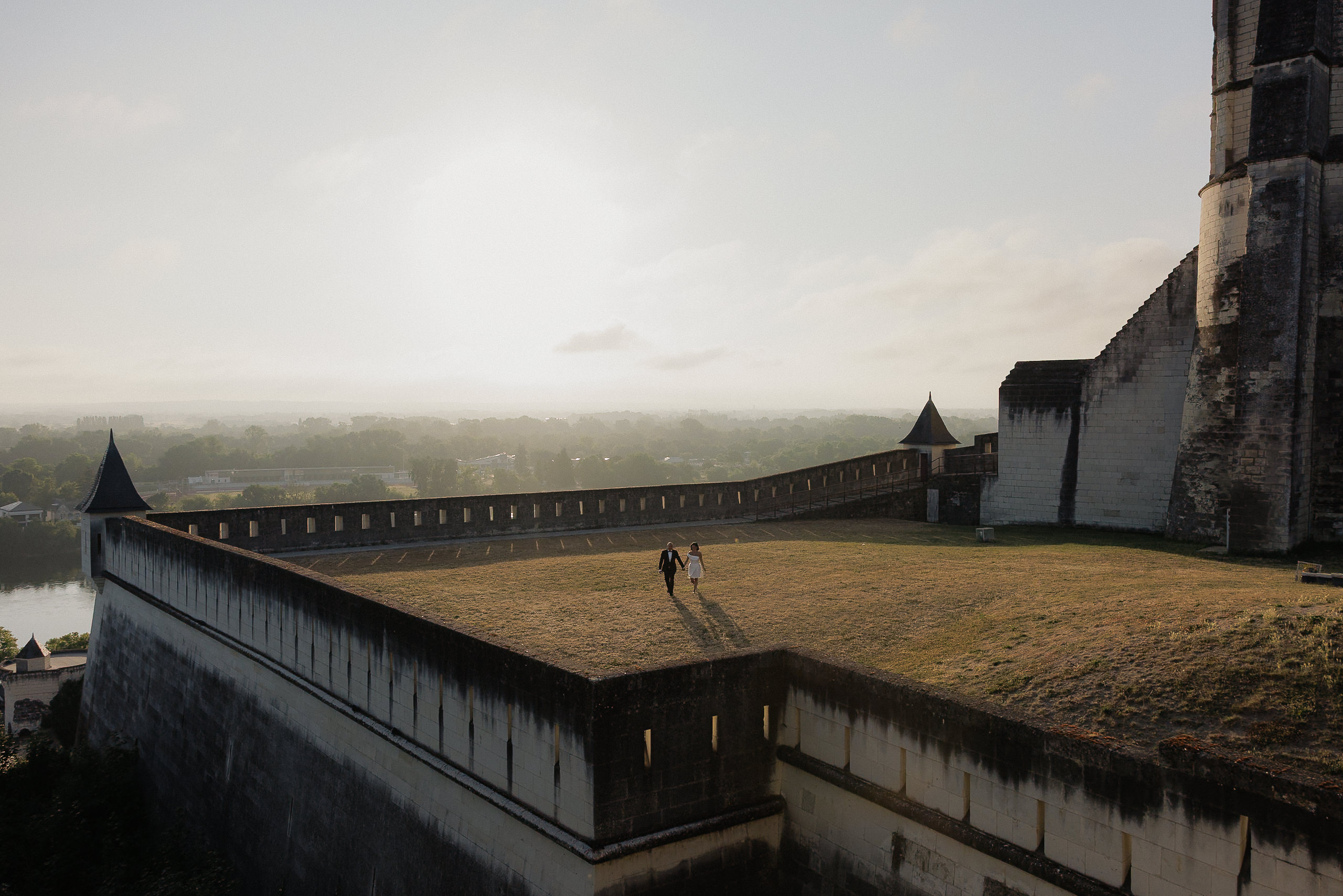 Two people walk hand in hand on a grassy courtyard at sunrise near a historic stone castle in Saumur, with soft light and haze.