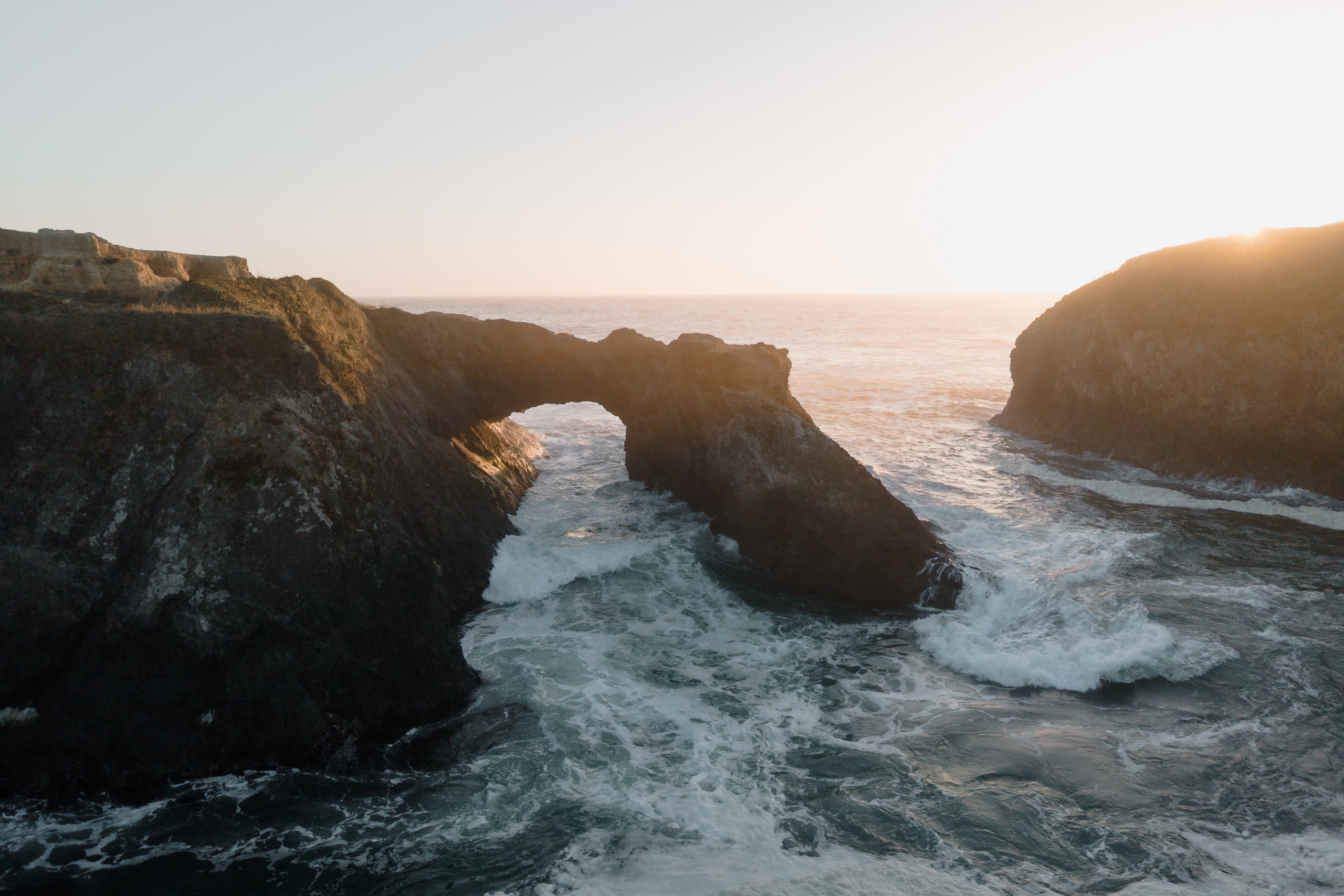 Capture Mendocinos coastal elegance: Waves crash through a rock arch at sunset, framed by rugged cliffs.