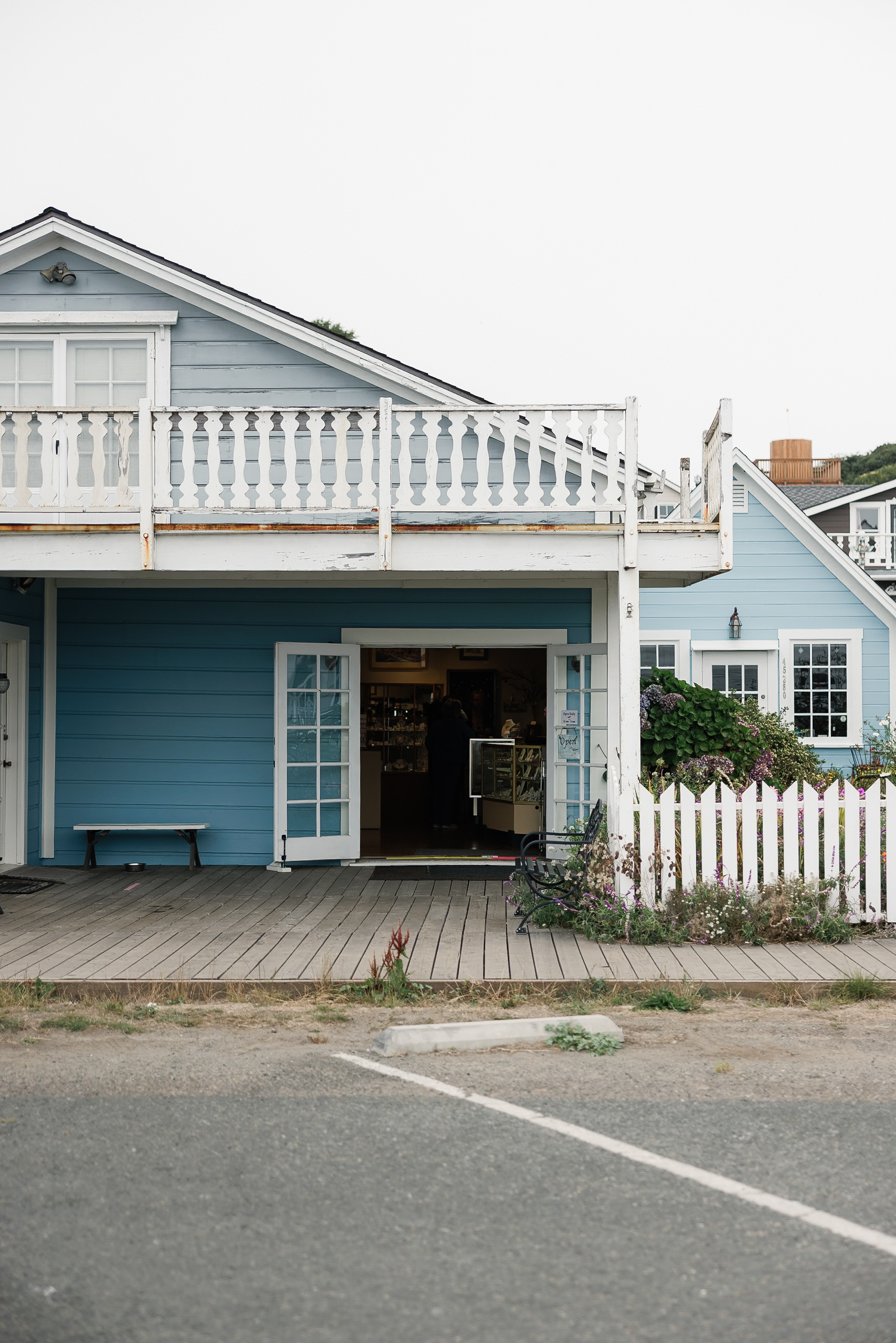 Elegant coastal cottage in Mendocino: blue facade, white trim, picket fence, purple blooms. Perfect wedding venue backdrop.
