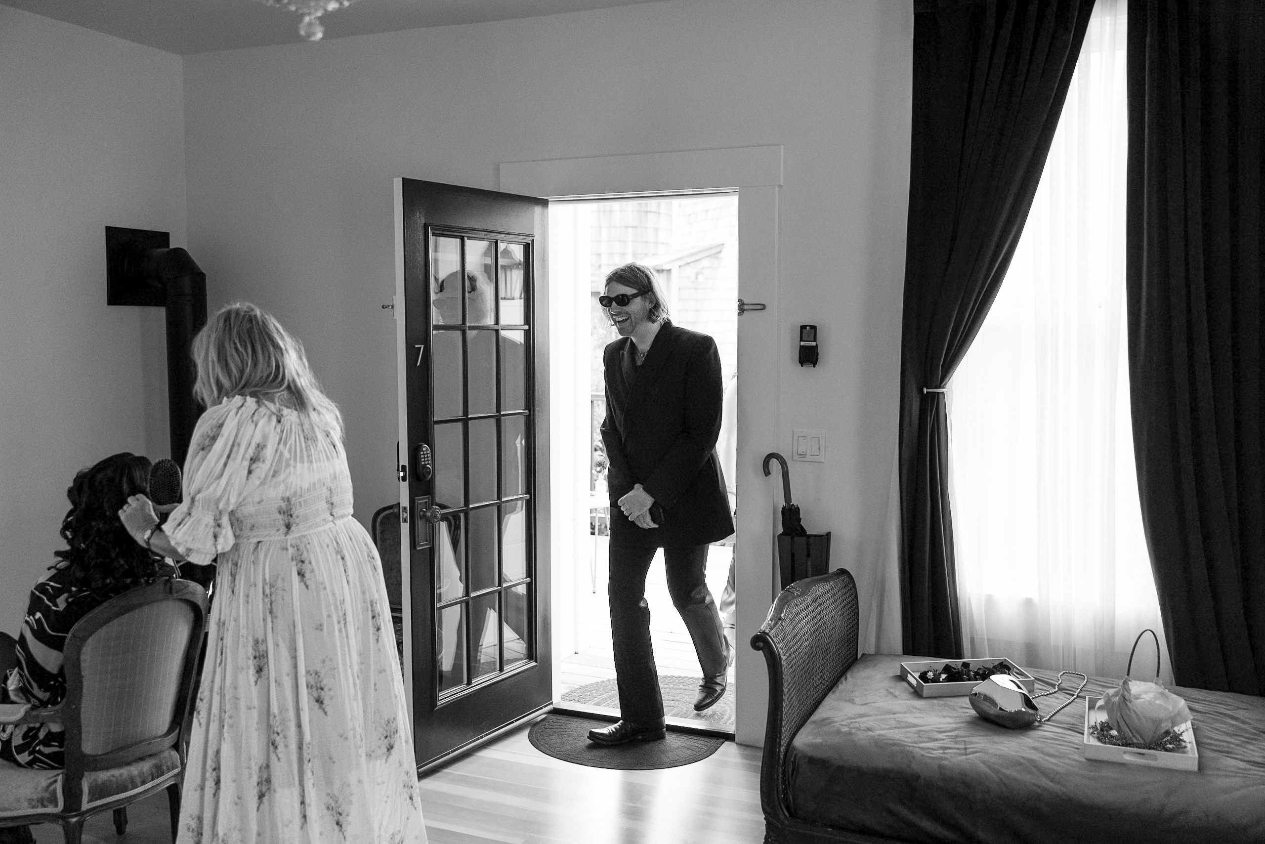 Mendocino Wedding: Man in Sunglasses Enters Sunlit Room with Daybed, Woman in Floral Dress Assists Seated Guest.
