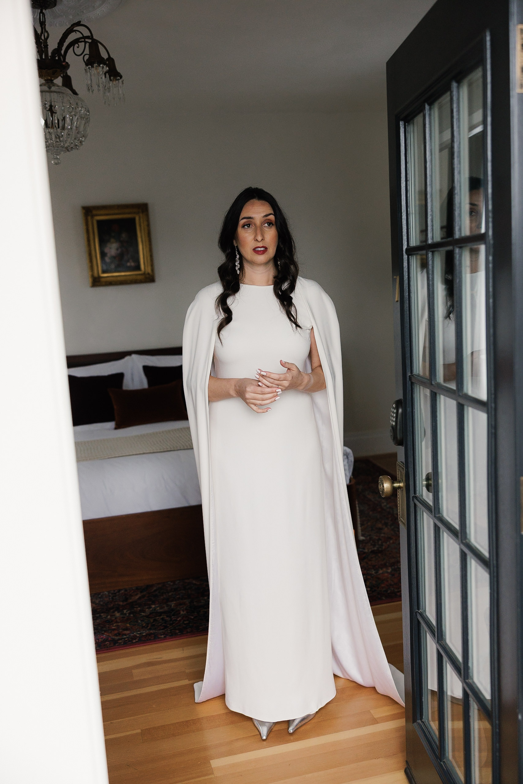 Elegant Mendocino bride: White gown with cape, flowing hair, chic bedroom; chandelier & framed art backdrop.