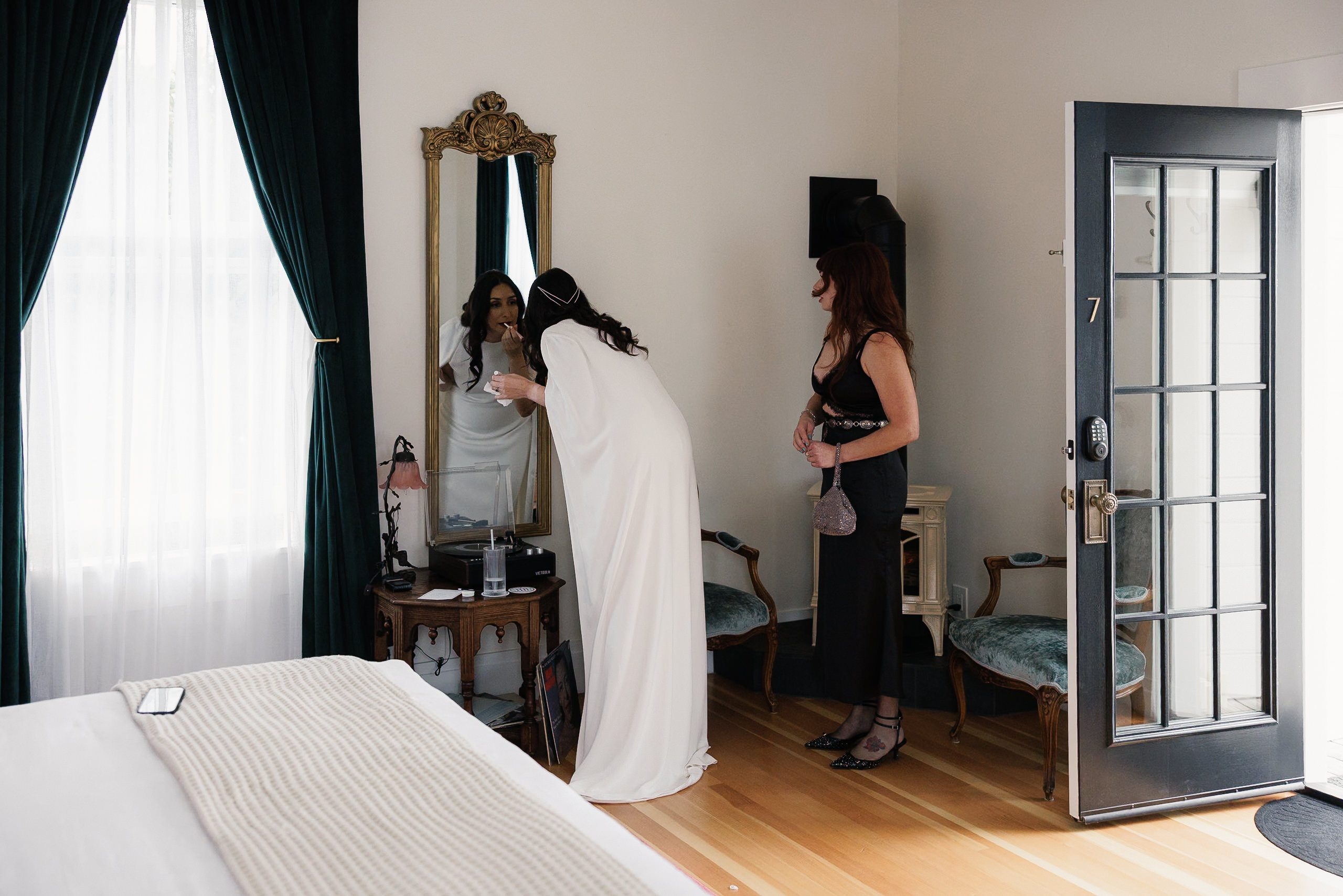 Elegant bride adjusts her hair in a Mendocino inn, with Room 7s curtains framing the scene.