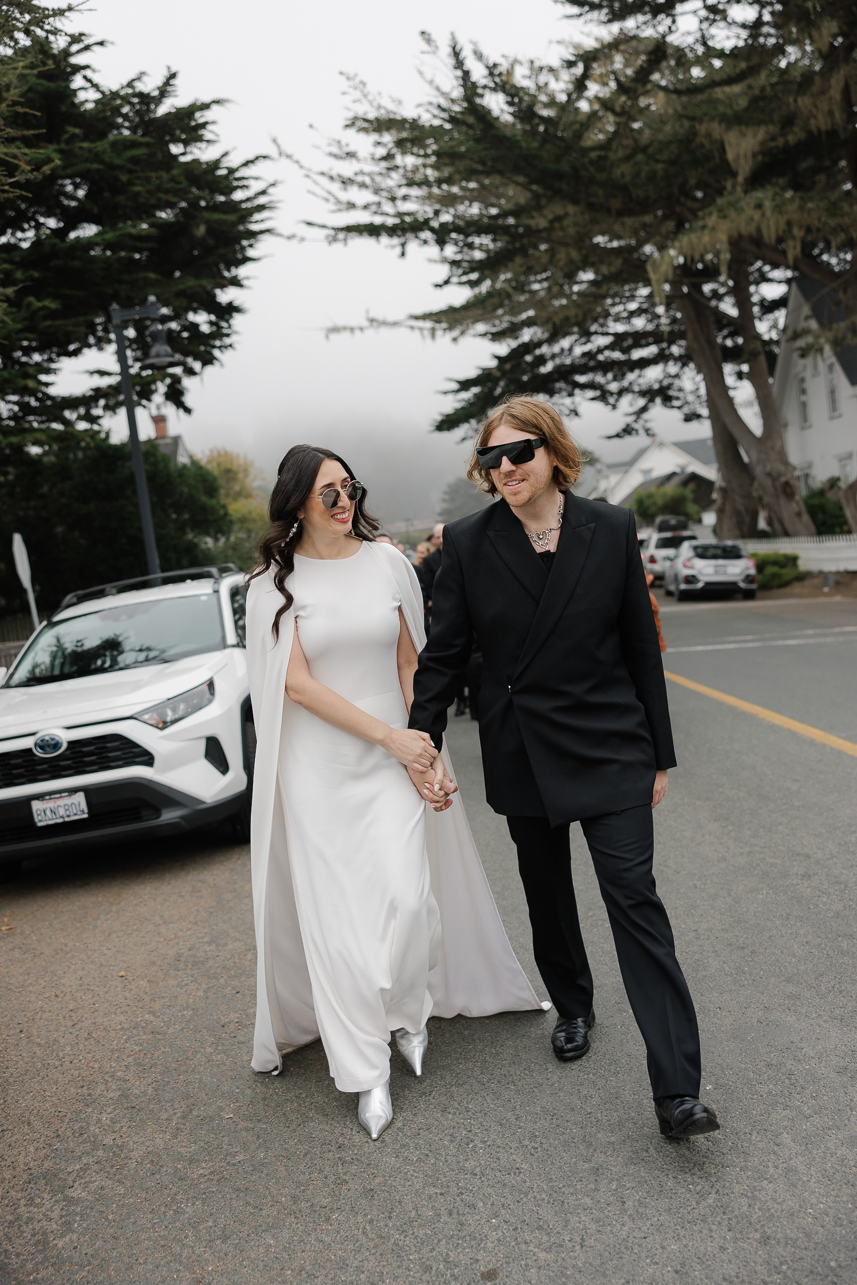 Mendocino Wedding Stroll: Bride and Groom in Stylish Attire Amid Misty, Tree-Lined Streetscape.