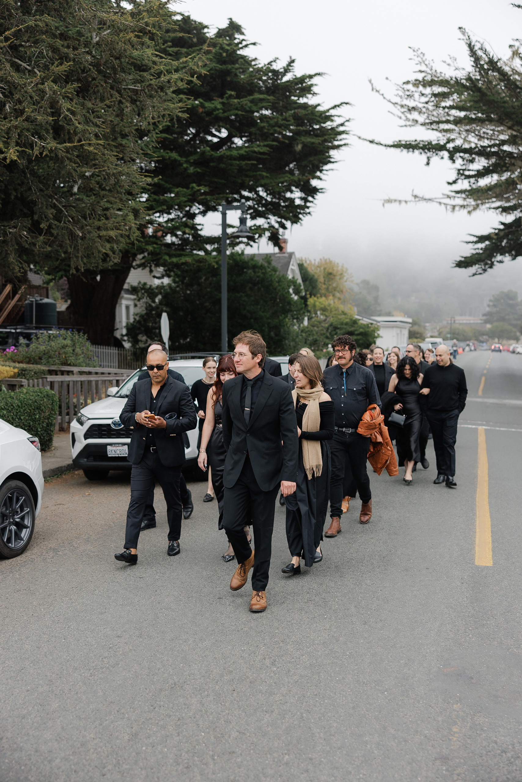 Elegant black-tie group strolls foggy Mendocino; leader checks phone. Lush trees and cars line street.
