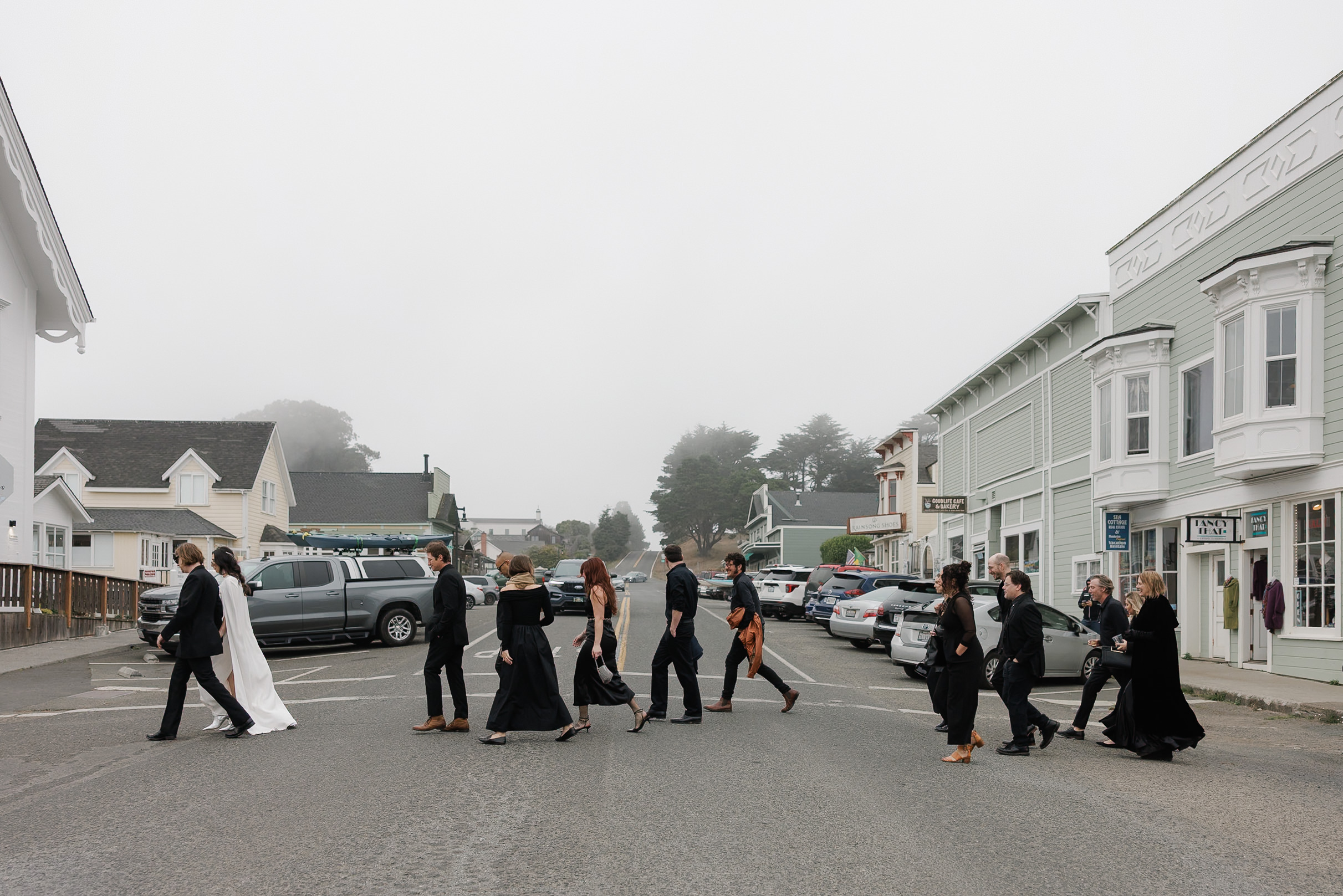 Elegant wedding party crossing foggy Mendocino street in serene, misty atmosphere.