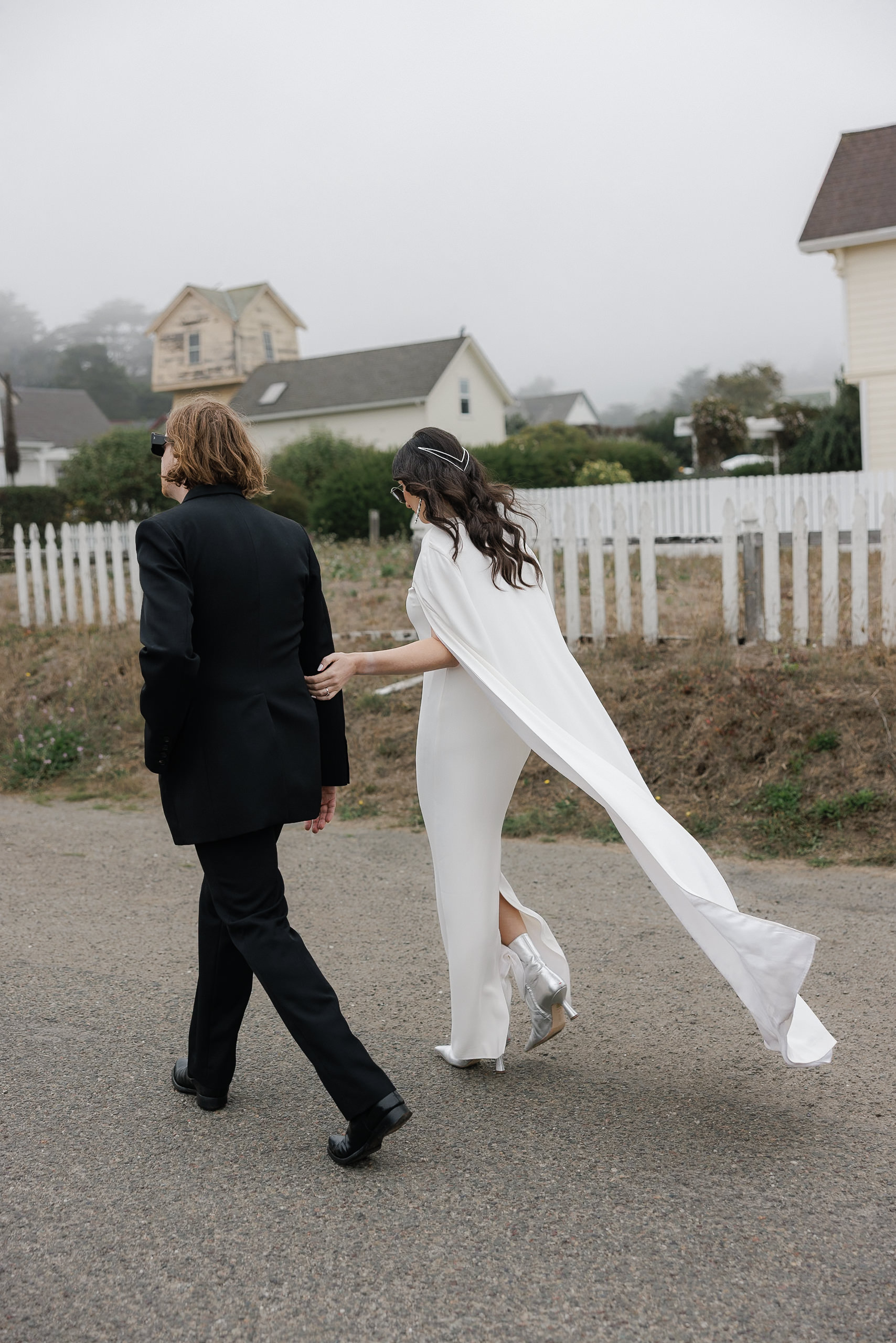Capture captivating wedding moments: bride in white cape dress and groom in black suit walking foggy Mendocino road.