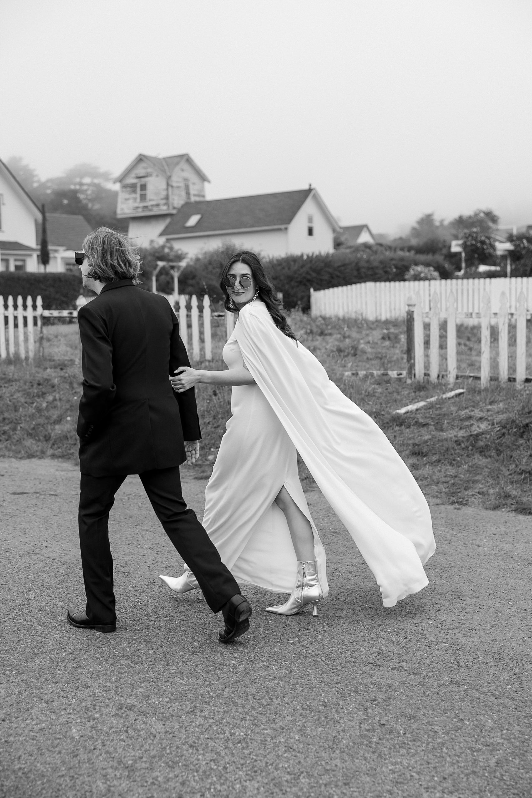 Elegant couple strolling in foggy Mendocino: flowing dress, black suit, white fences backdrop.