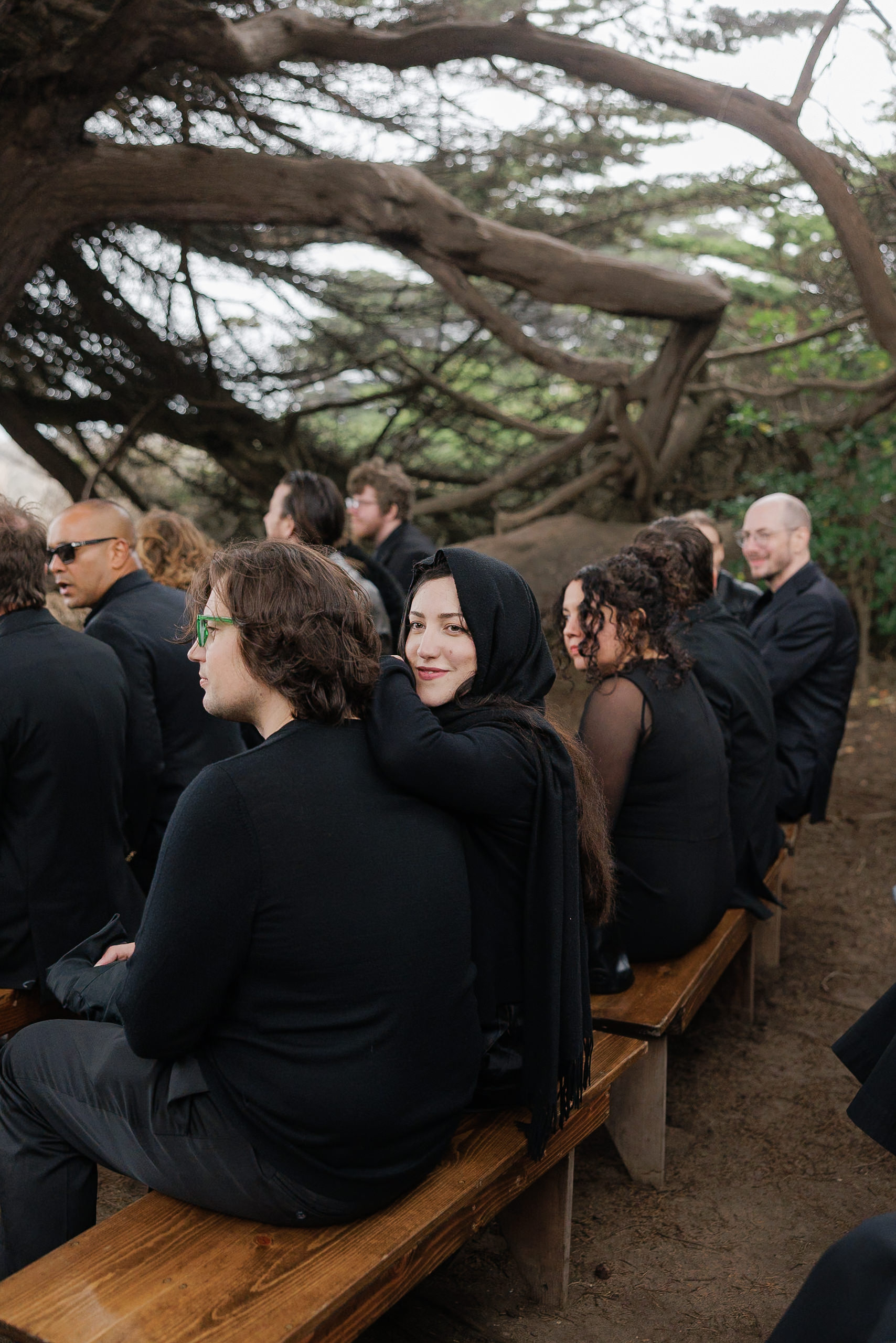 Mendocino outdoor wedding: guests in dark attire on wooden benches, under grand trees; a woman smiles warmly.