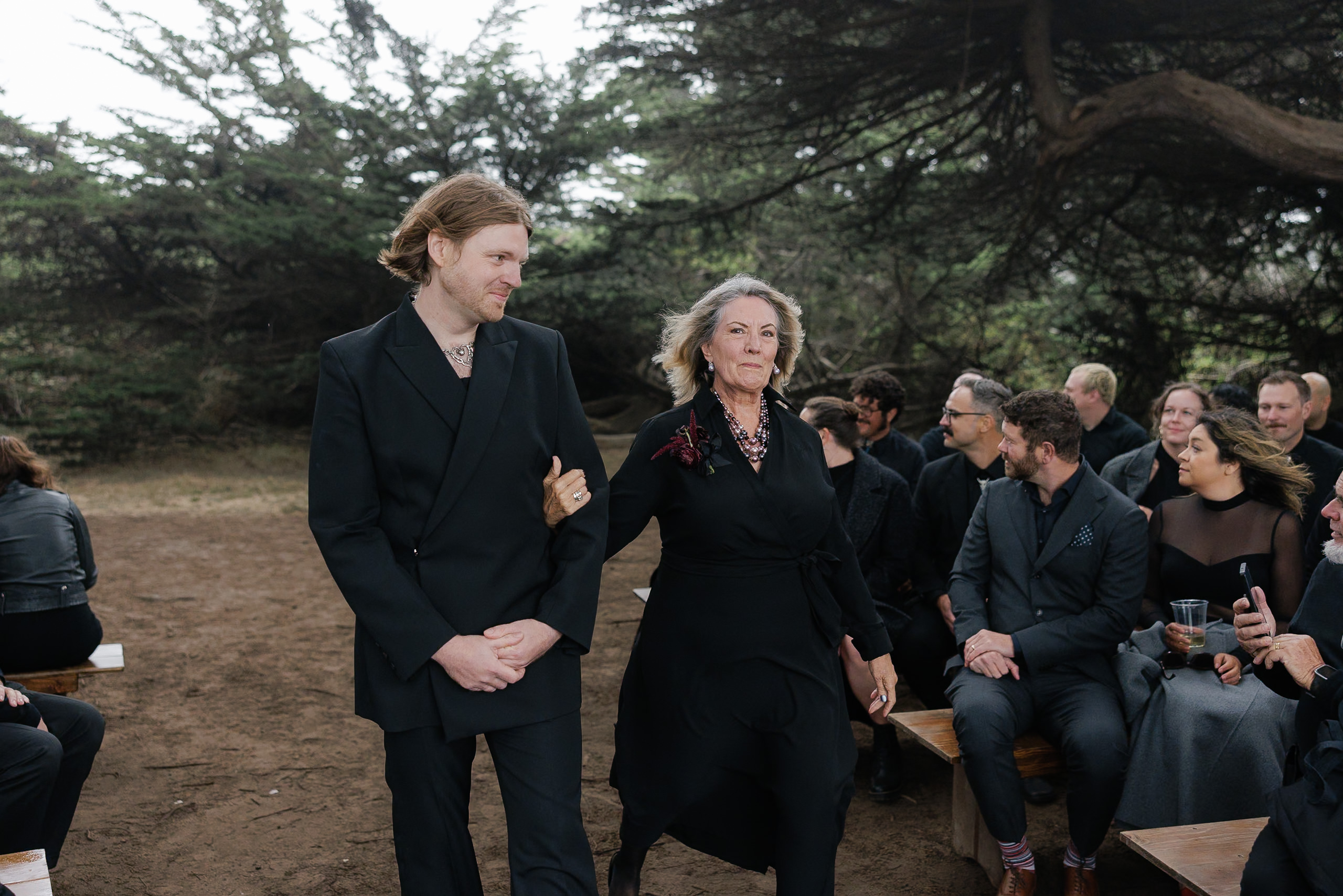 Elegant outdoor gathering in Mendocino: Stylishly dressed attendees observe amidst towering trees.