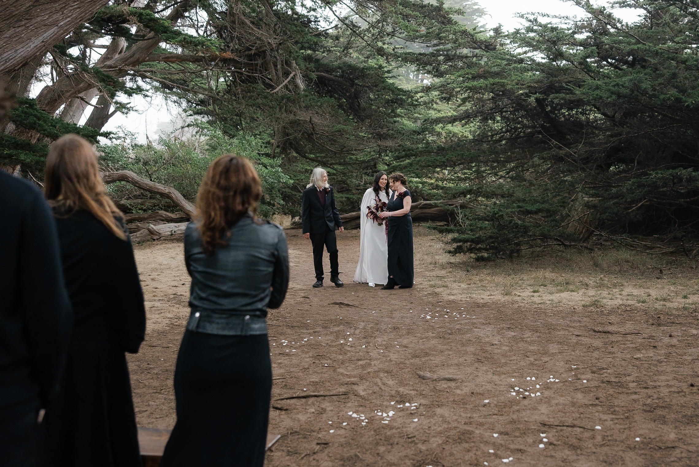 Elegant bride in white with purple flowers on Mendocino forest path; serene ceremony among tall trees.